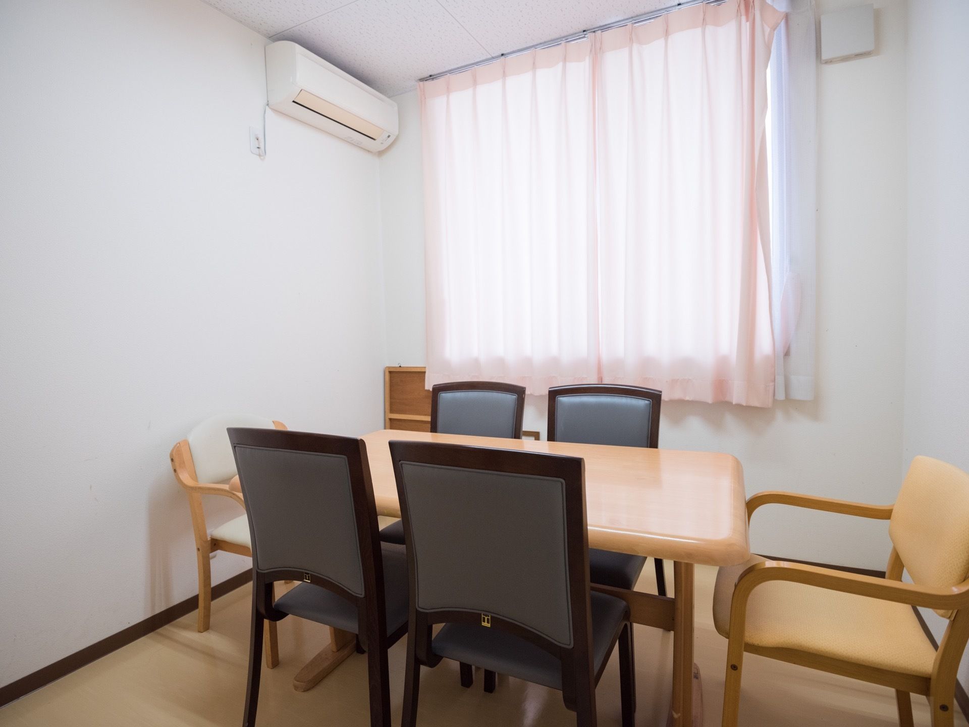 部屋の中央にある、薄茶色のテーブルを囲むように椅子が６脚置かれている。窓にはカーテンがかけられ、エアコンが設置されている。