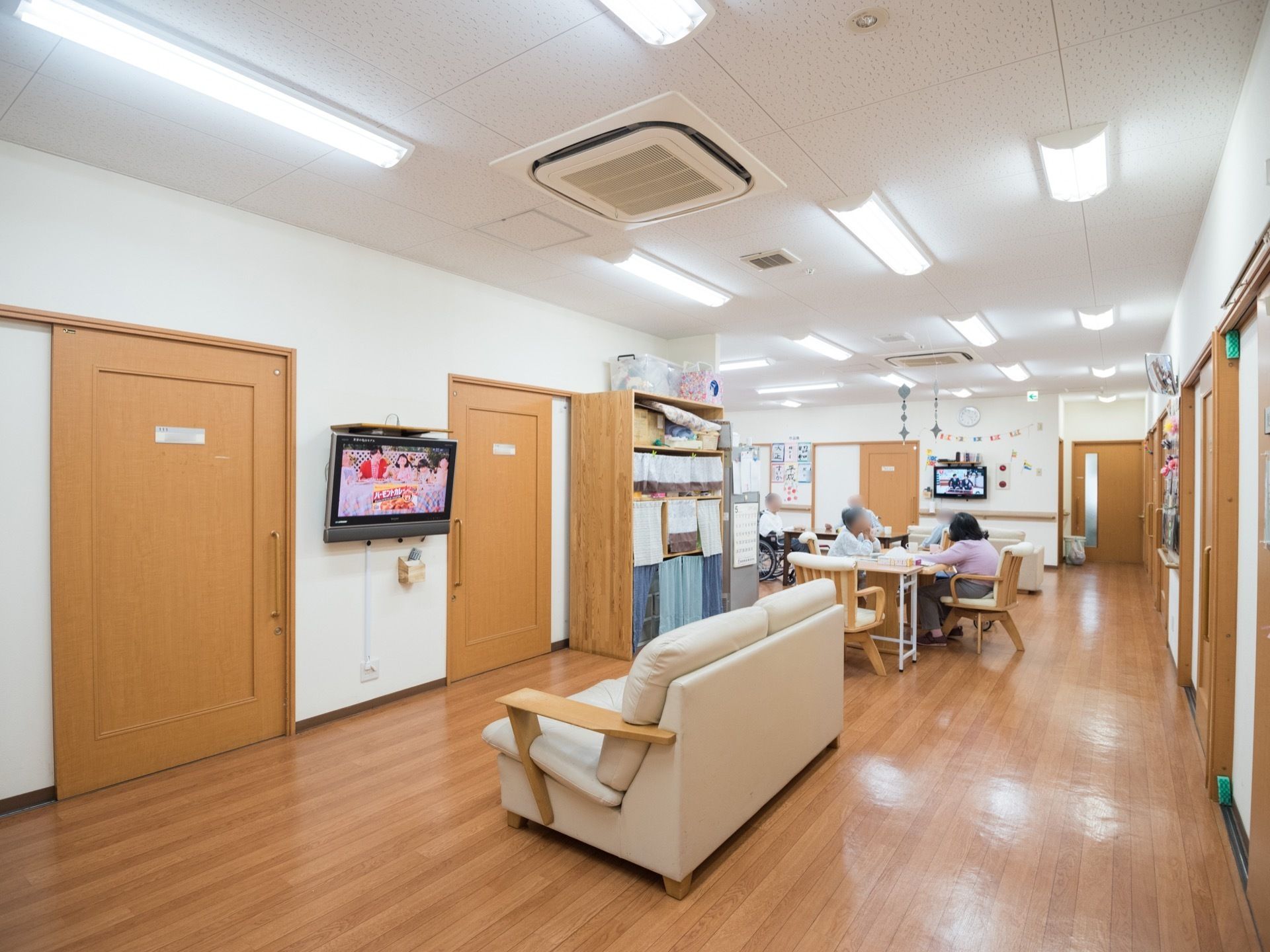 中央に白い大きなソファが置かれており、その前には壁掛けのテレビが設置されている。また、引き戸式の部屋への入口扉も見える。