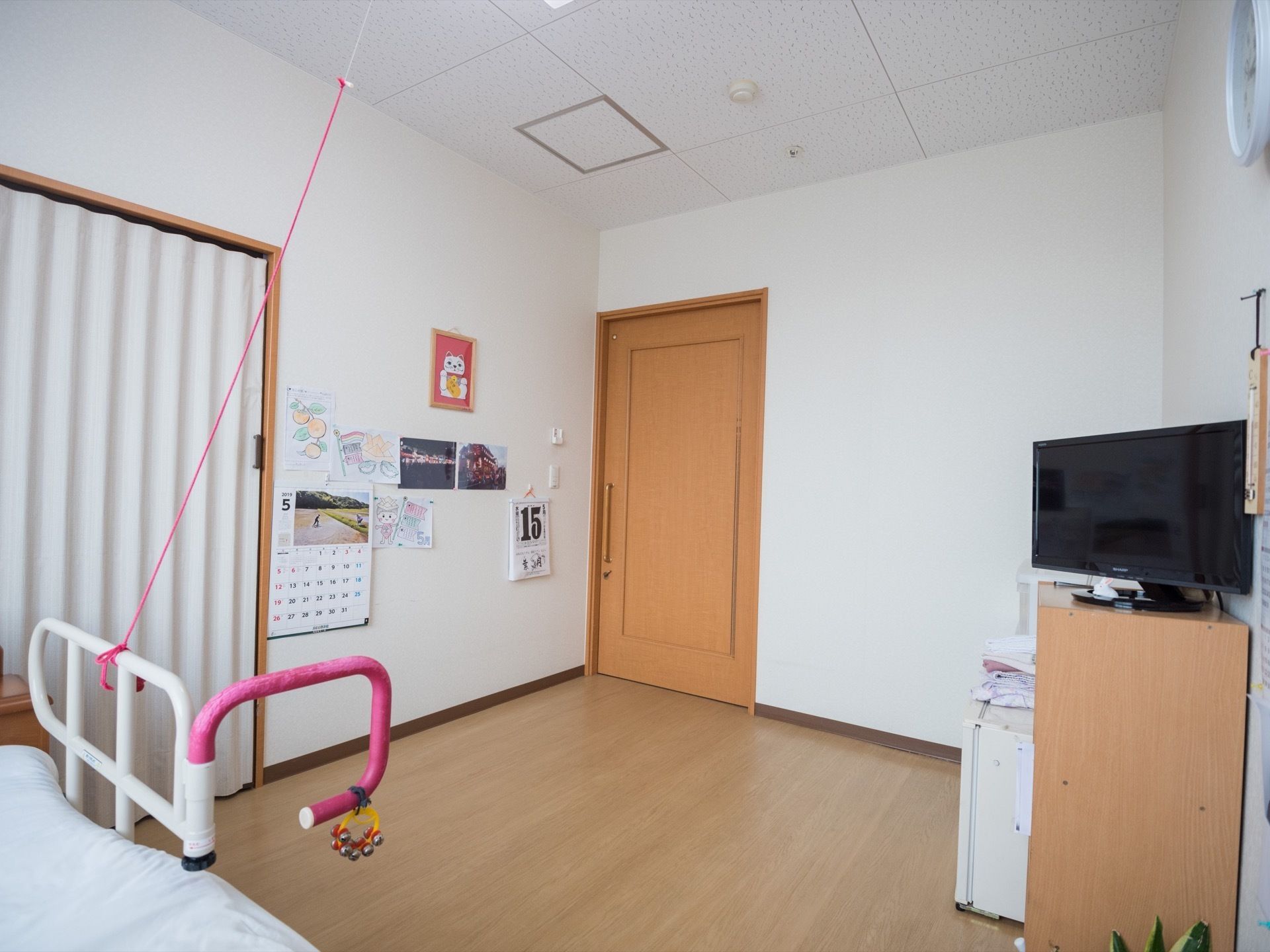 白い壁の居室である。アコーディオンカーテンで仕切られた場所があり、その横の壁には、カレンダーや写真が貼られている。