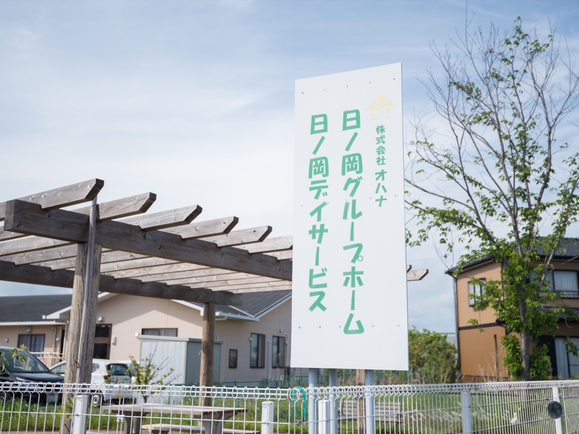 白い金属製のフェンスの上に、施設名が書かれた看板が立っている。施設の駐車場には、車が駐車しており、近くの住宅も見える。