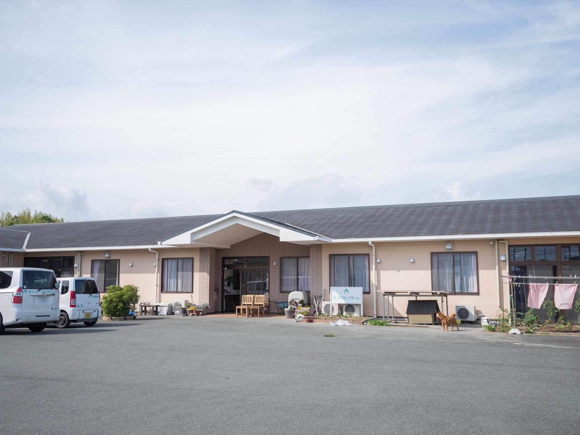 平屋建てのグループホーム。建物前にはアスファルトで舗装された駐車場が設けられており、玄関横には犬小屋がある。