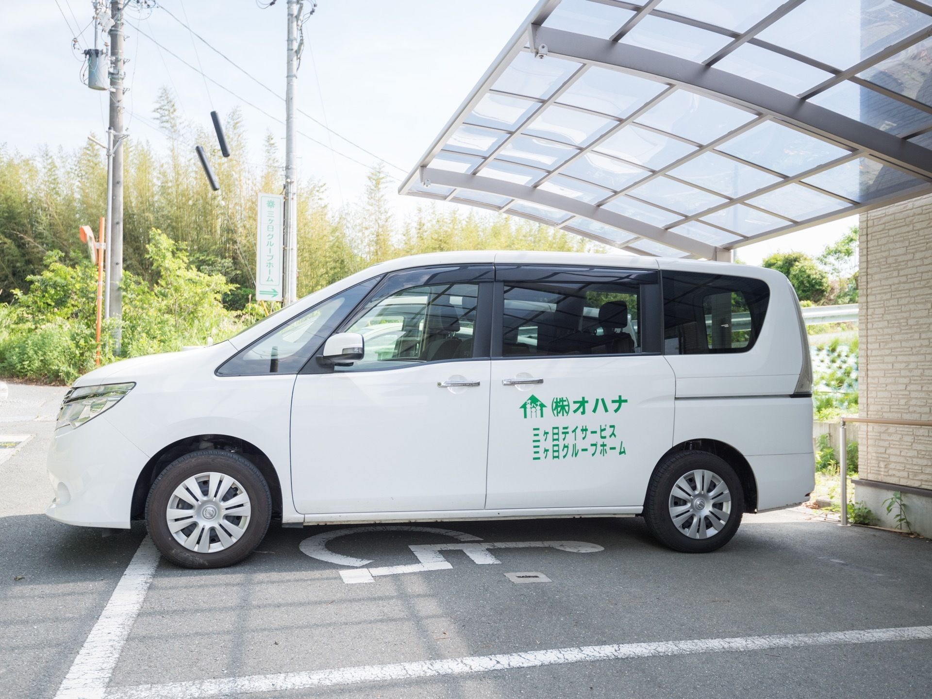 施設駐車場に送迎車が停められている。白い大きなワゴンタイプの送迎車で複数人が乗れるようになっている。ドアには施設名が書かれている。