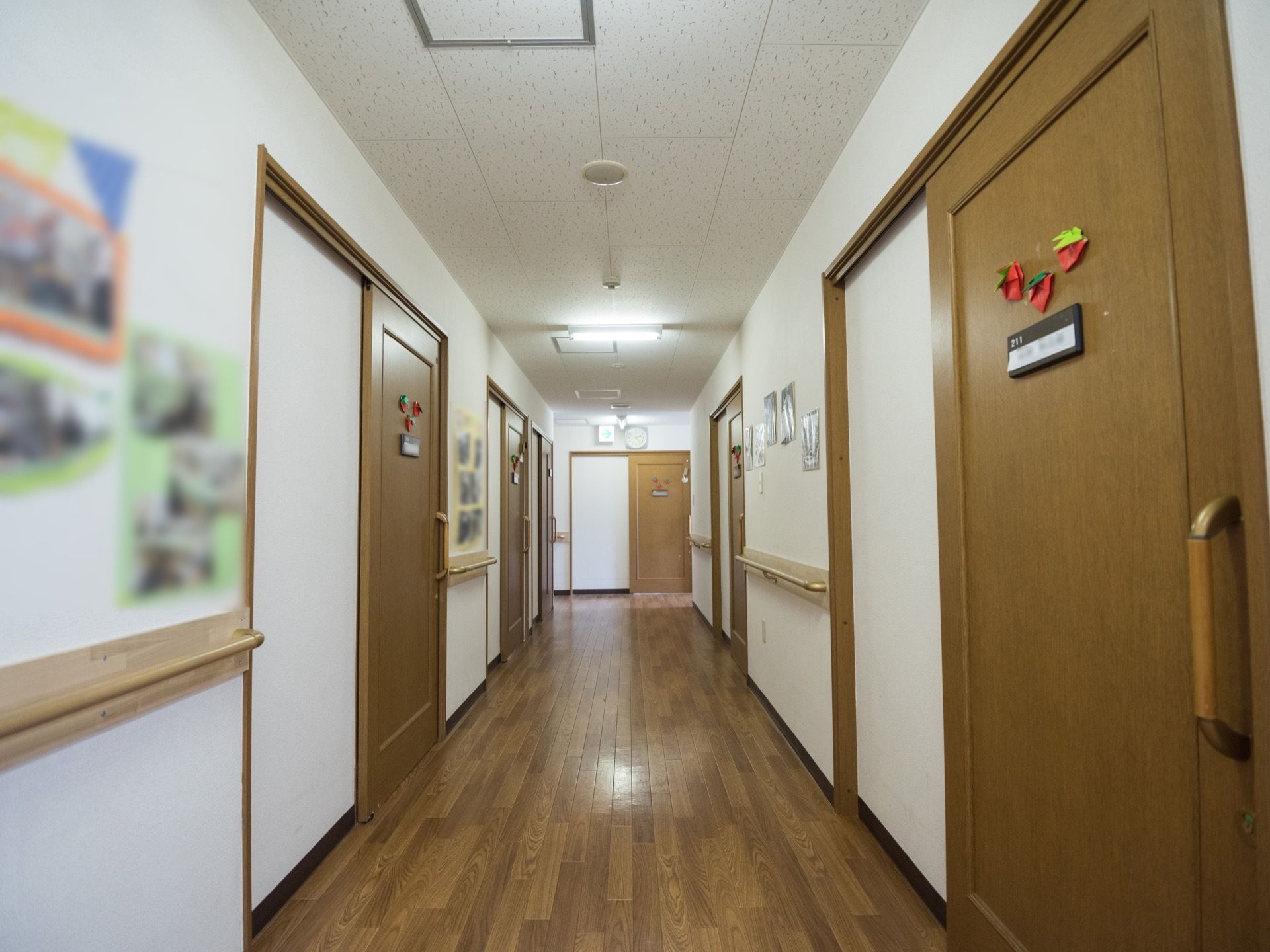 木目調の床が落ち着いた雰囲気を出している。両脇に部屋が並んでいて、壁沿いには手すりが設置され、壁には複数の写真が飾られている。