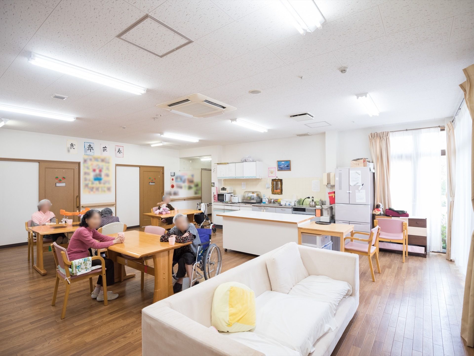 広く、明るい雰囲気の室内である。テーブルや椅子、ソファーなどが置かれていて、奥にはキッチンも見える。