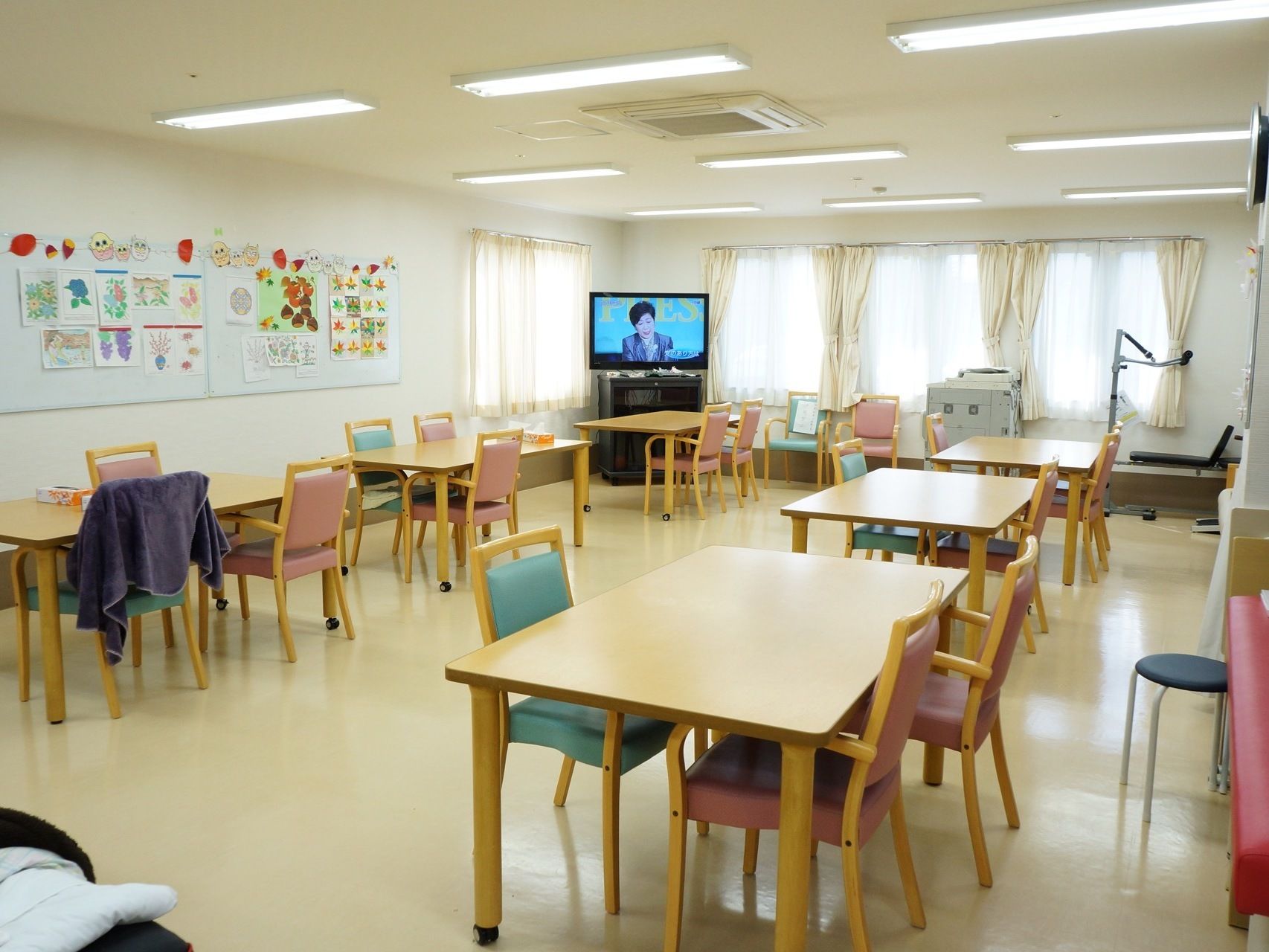 4人がけのテーブルがそれぞれ独立して6卓ある。壁にはご利用者様の作品が飾られ部屋の角にテレビがある。腰高窓がある。