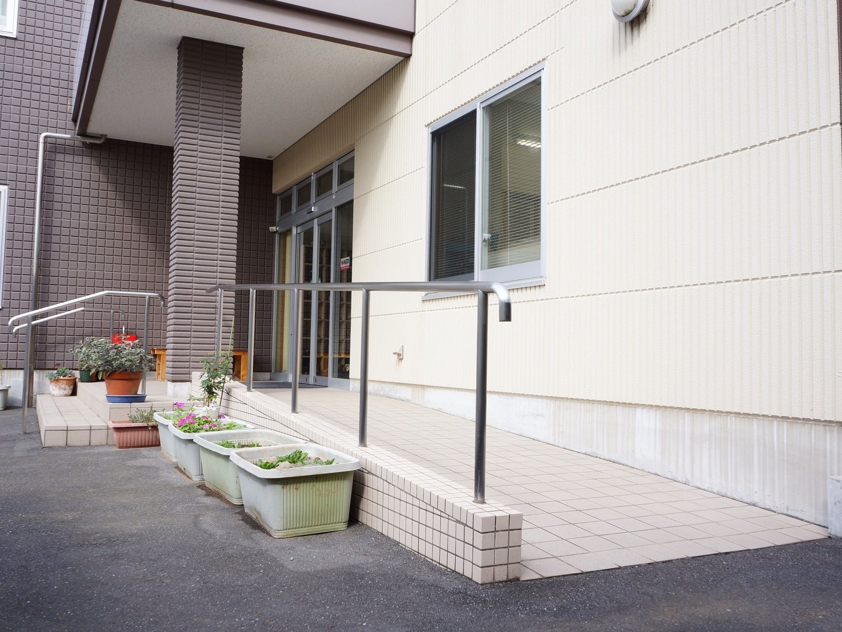 建物玄関正面に低めの2段の階段がある。玄関から建物に沿って手すり付きのスロープがある。階段には両側に手すりがある。