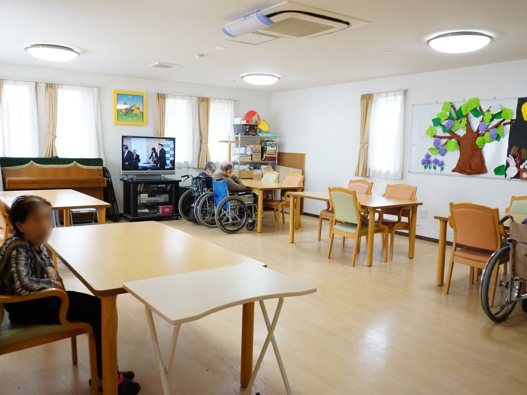 共有スペースには４人の入居者様がおり、３人は車イス、もう１人は施設のイスにすわっている。テレビは電源がついていて、番組が映っている。