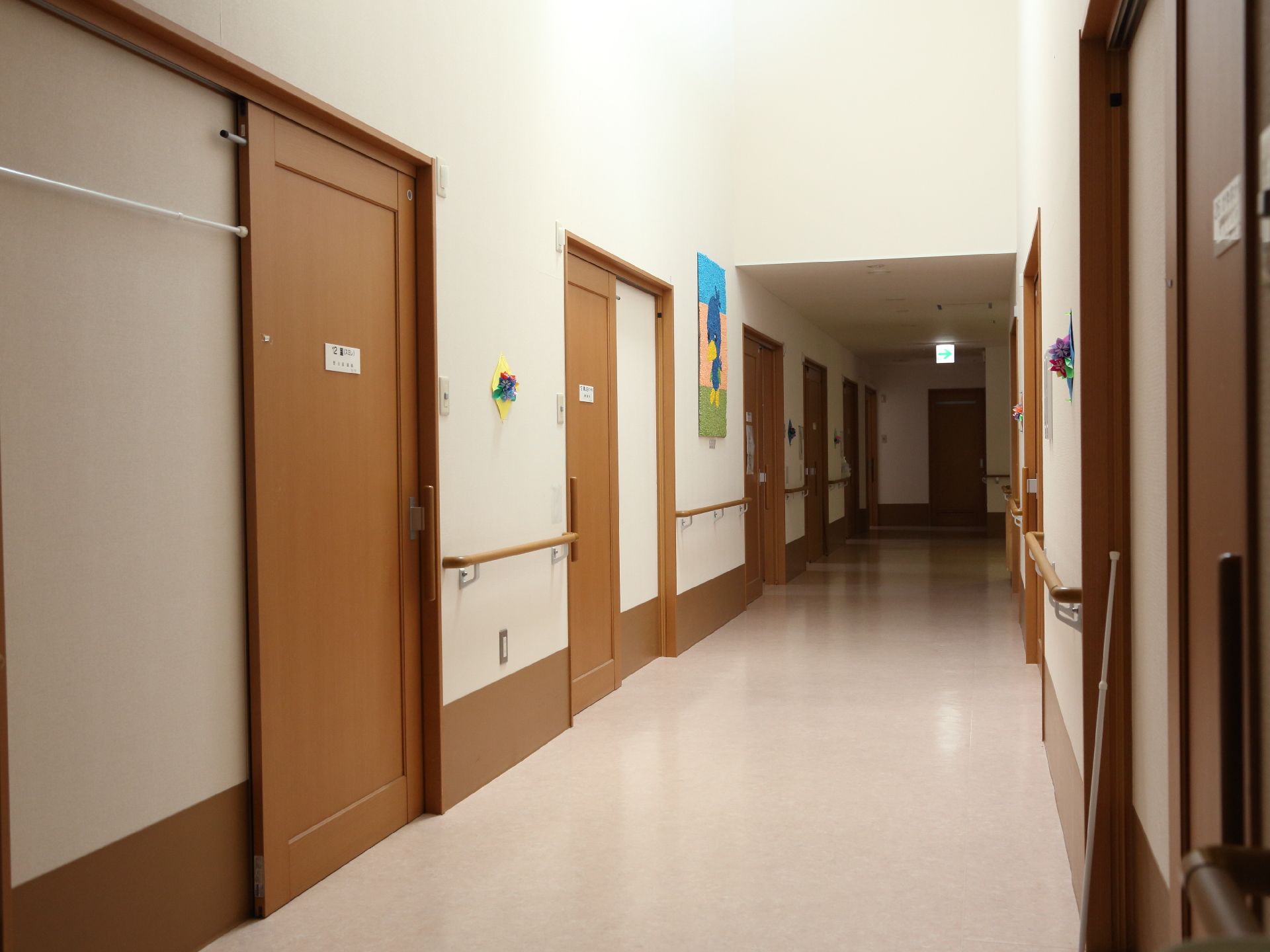 車椅子がすれ違える広さがある。壁には手摺、絵や折り紙で作られた飾りがある。奥には非常口が設けられている。