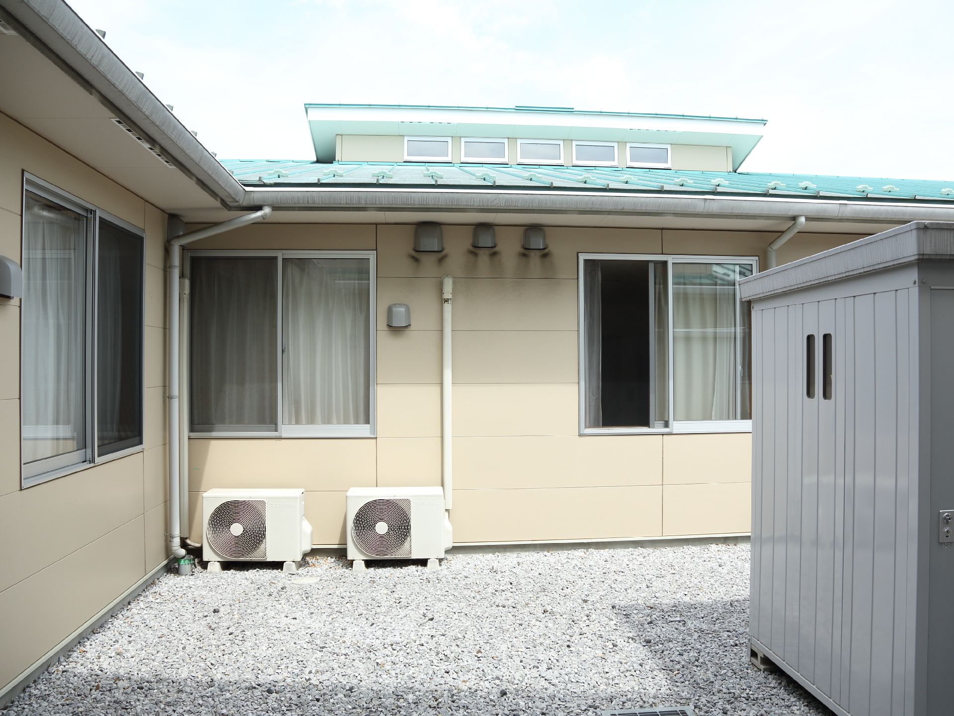 クリーム色の外壁と青い屋根の平屋づくりの建物である。窓の外にはエアコンの室外機が置かれ、砕石が敷いてある。