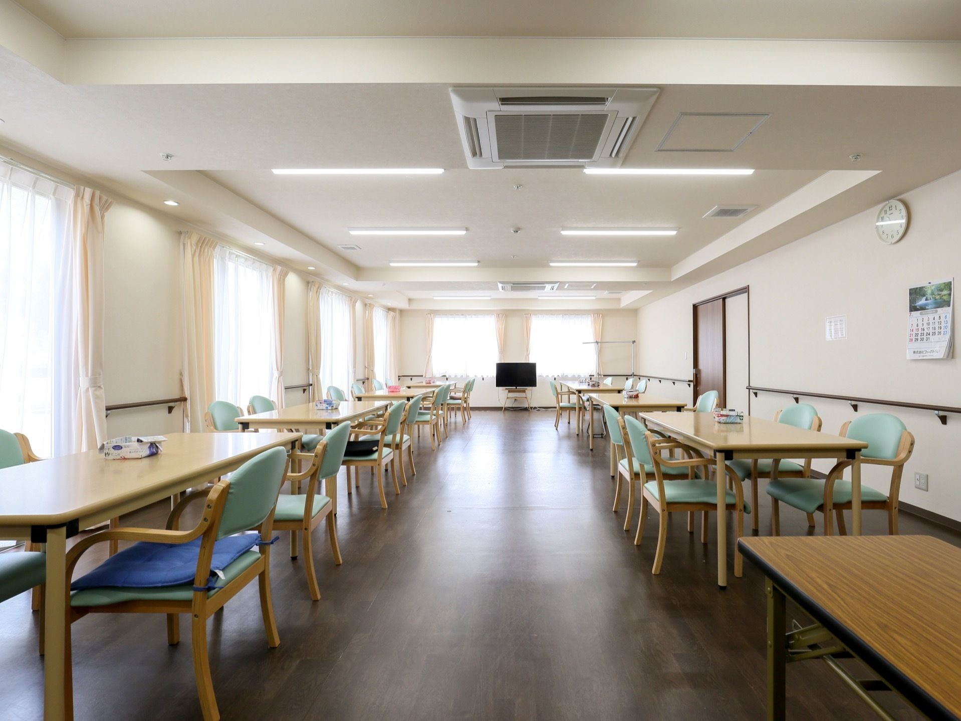 多くのテーブルが並べられている食堂。大きく取られた窓により日当たりを確保している。部屋の一角にはテレビがある。