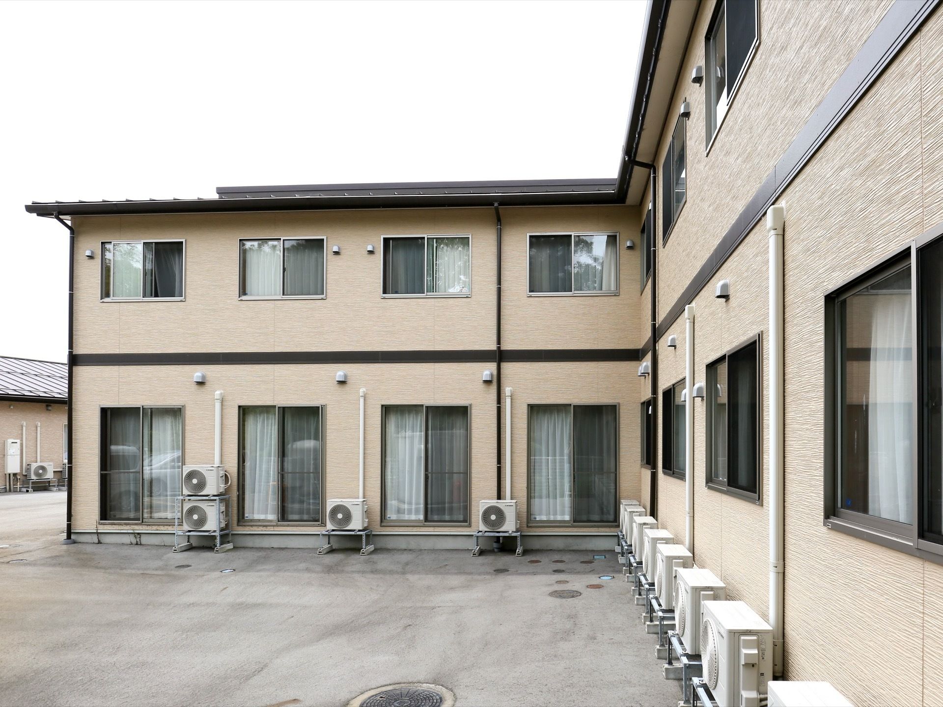 建物は２階建て構造となっている。クリーム色の外壁には、居室や各設備用のエアコン室外機が取り付けられている。