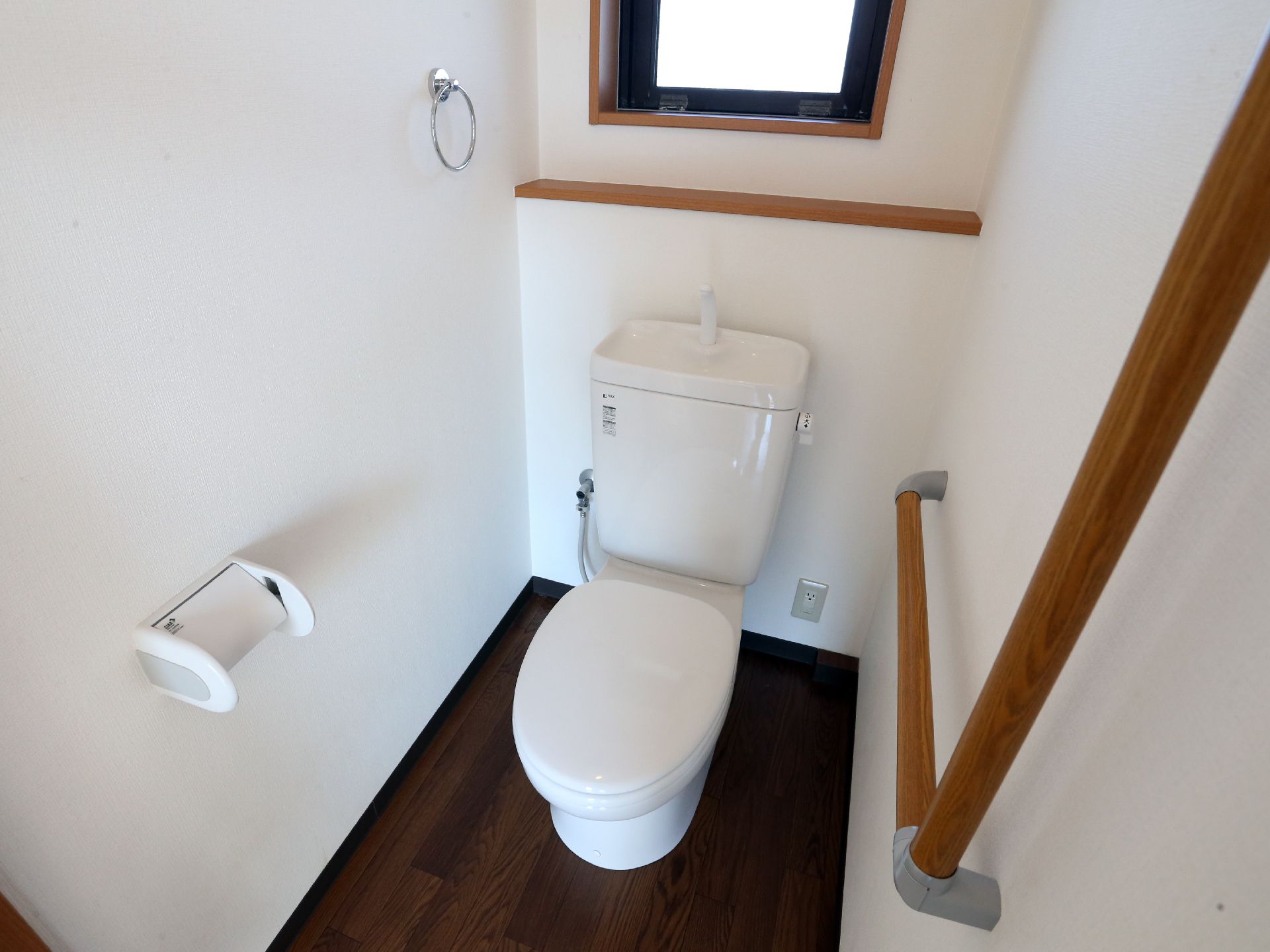 窓がついたトイレには洋式便器がある。木目のフローリングフロアの清潔感ある空間である。壁には手すりがついている。