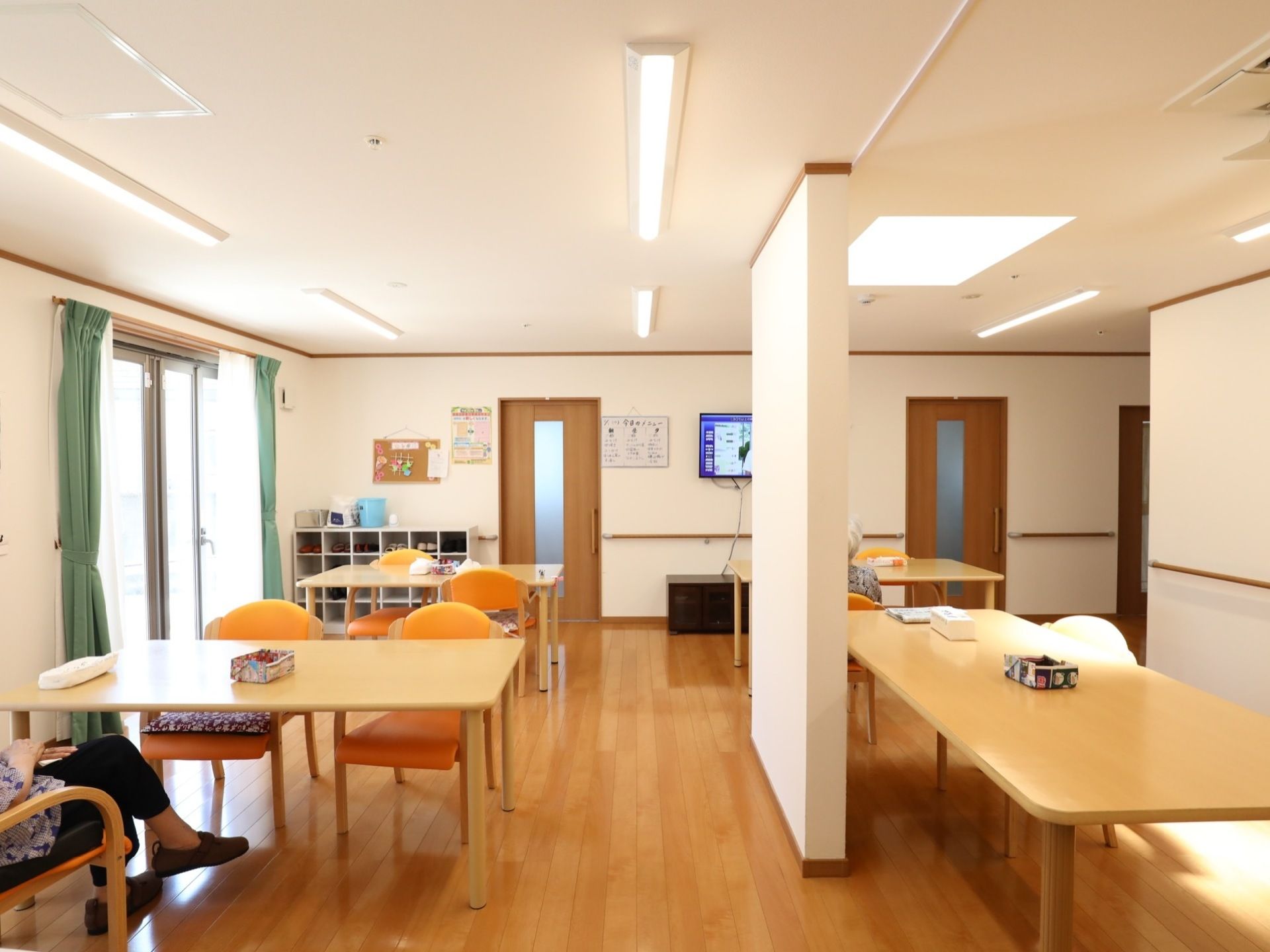 蛍光灯が複数設置されている明るいリビングで、入居者様が椅子に座ってテレビを観ている。窓際の壁には靴箱が置かれている。
