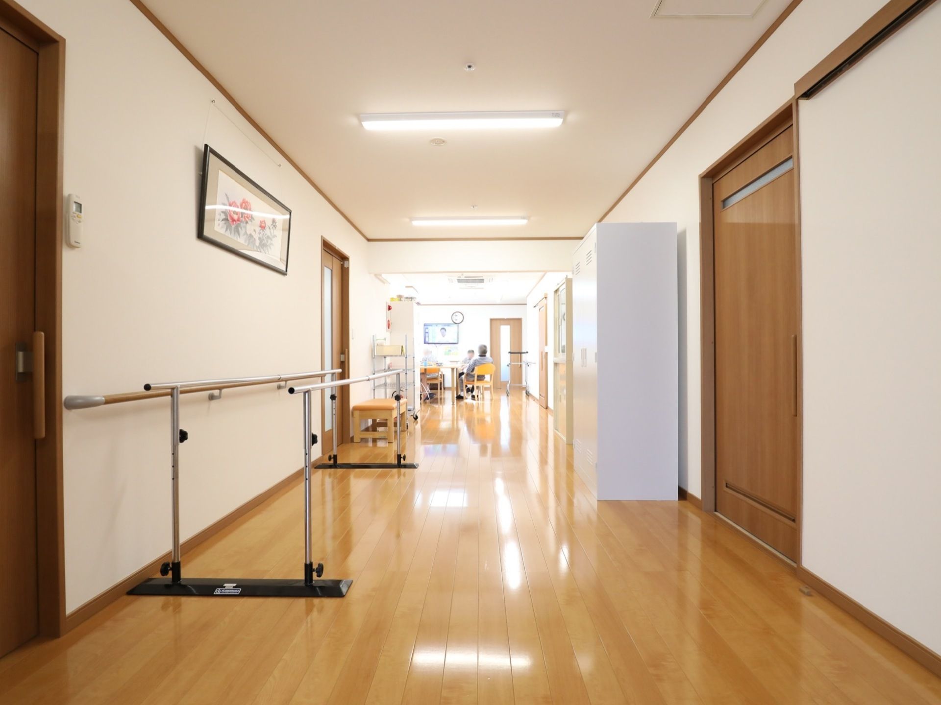 壁に手すりやリモコン、額に入れられた絵が掛けられている。奥にはリビングがあり、入居者様が座っているのが見える。