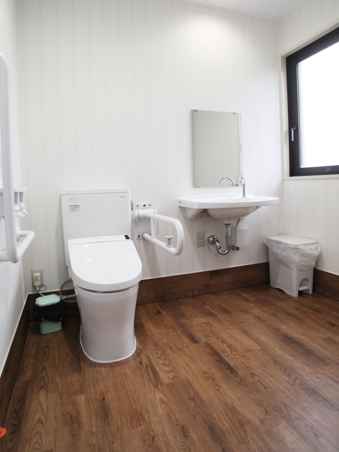 大きな窓と白い壁面で明るいトイレには手すりが左右にあり、洗面台もある。車椅子でも十分入れるスペースがある。