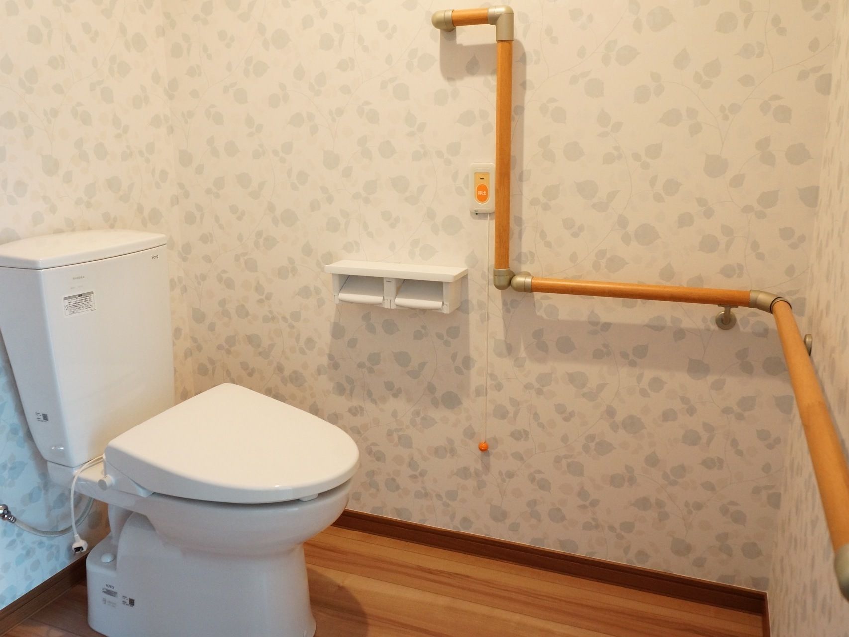 トイレ内の２方向の壁には木製の手すりがある。座って左側の壁には小物棚付きのペーパーホルダーとナースコールがある。