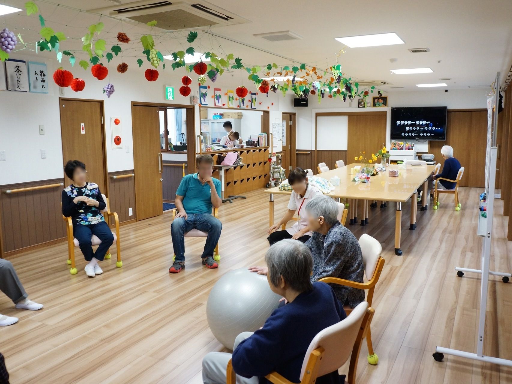 ボールを用いたアクティビティを行っている。共有スペースのテレビは付いていて、テレビ鑑賞をしている利用者様もいる。
