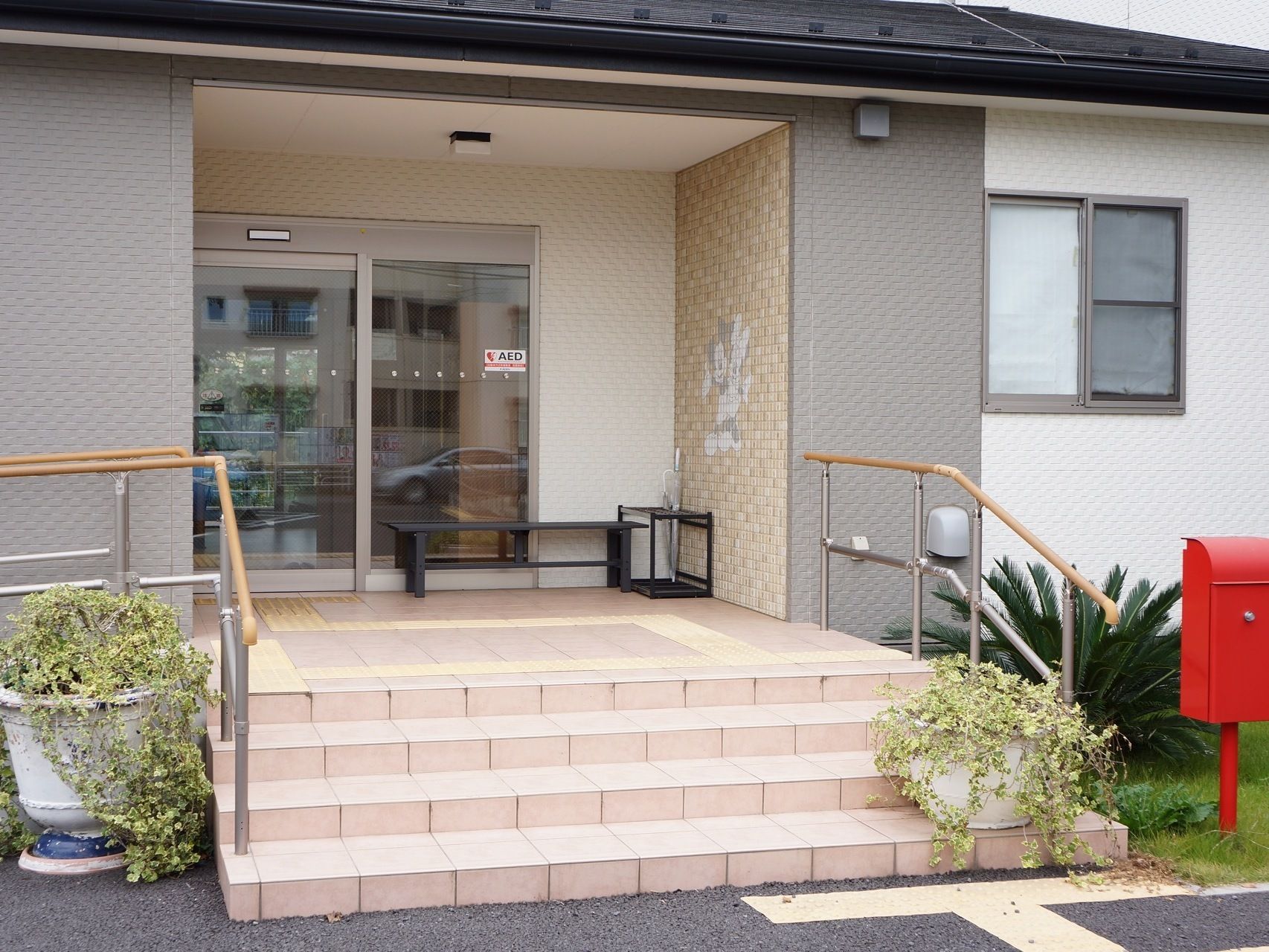 正面玄関の外にベンチが置かれている。上には屋根になる部分がある。ベンチの隣には傘立てが置かれている。