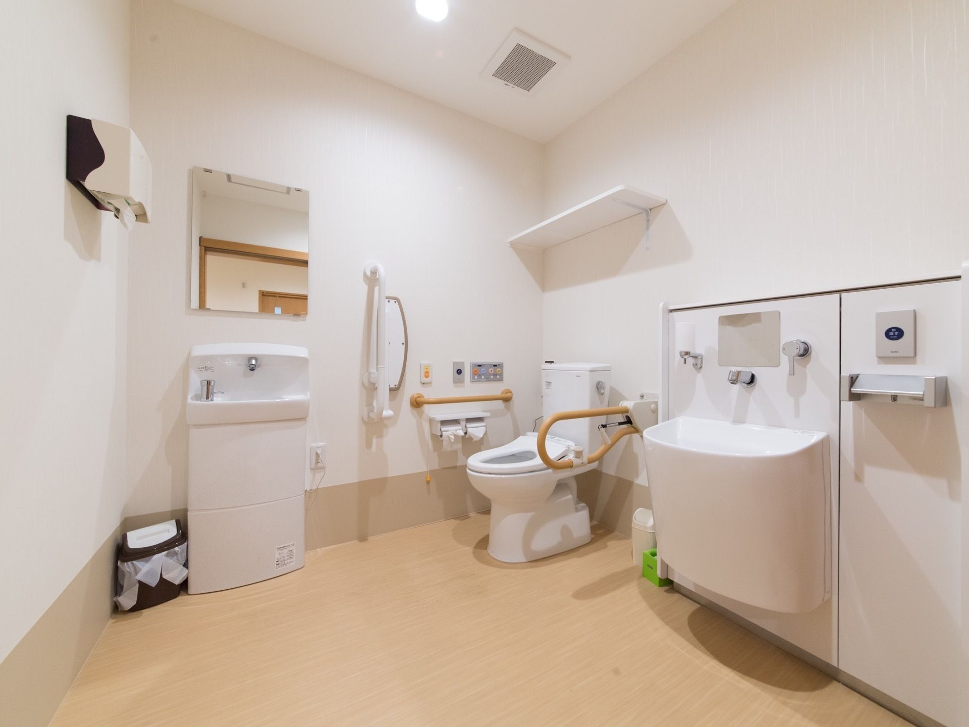 オストメイト対応トイレを備えた個室トイレである。洗面台やナースコール、跳ね上げ式の手すりなどが設置されている。