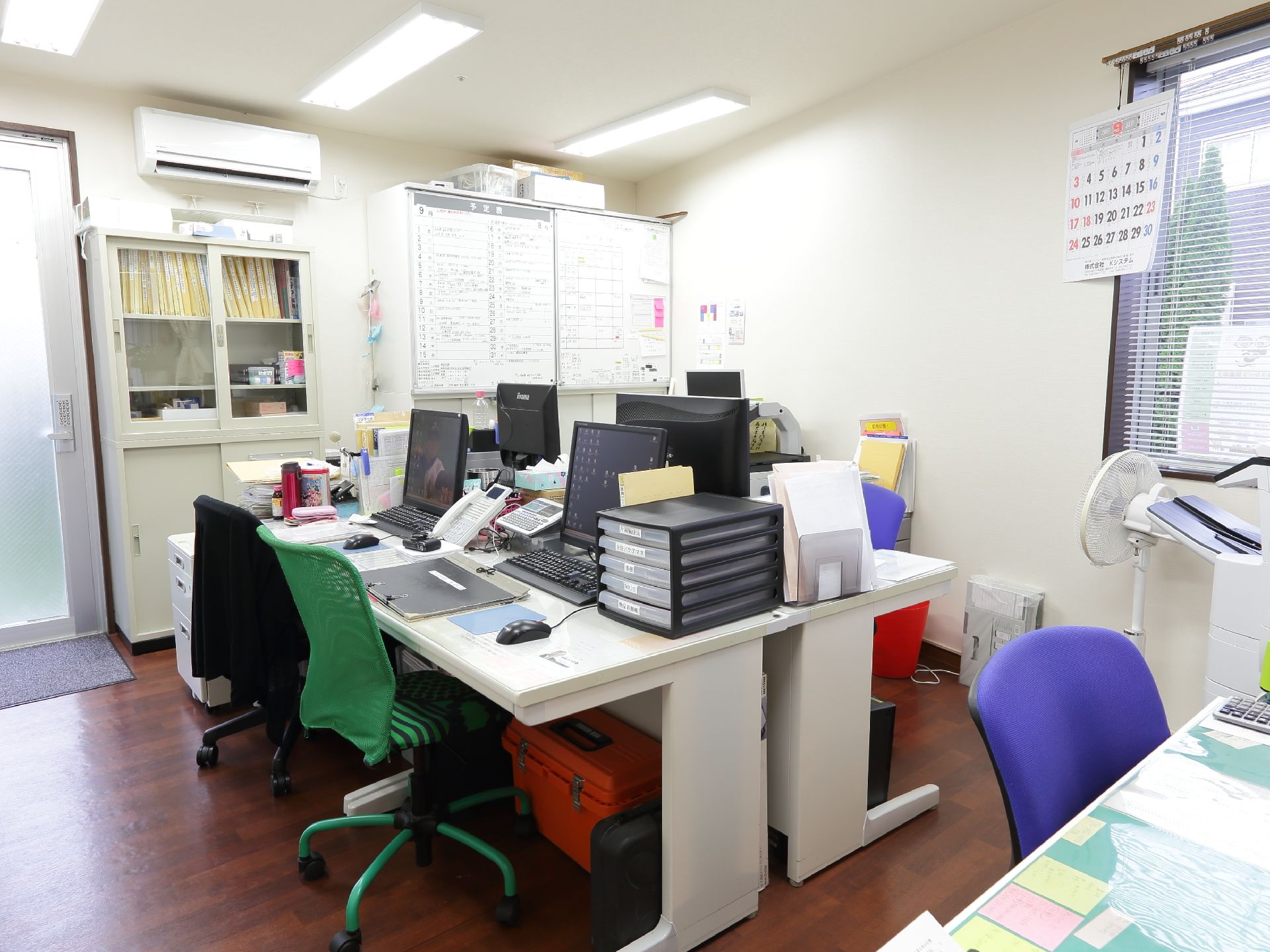 事務室の壁には空調設備があり、さまざまな書類が並べらた棚が置かれている。また大きな窓にはブラインドが取り付けられている。