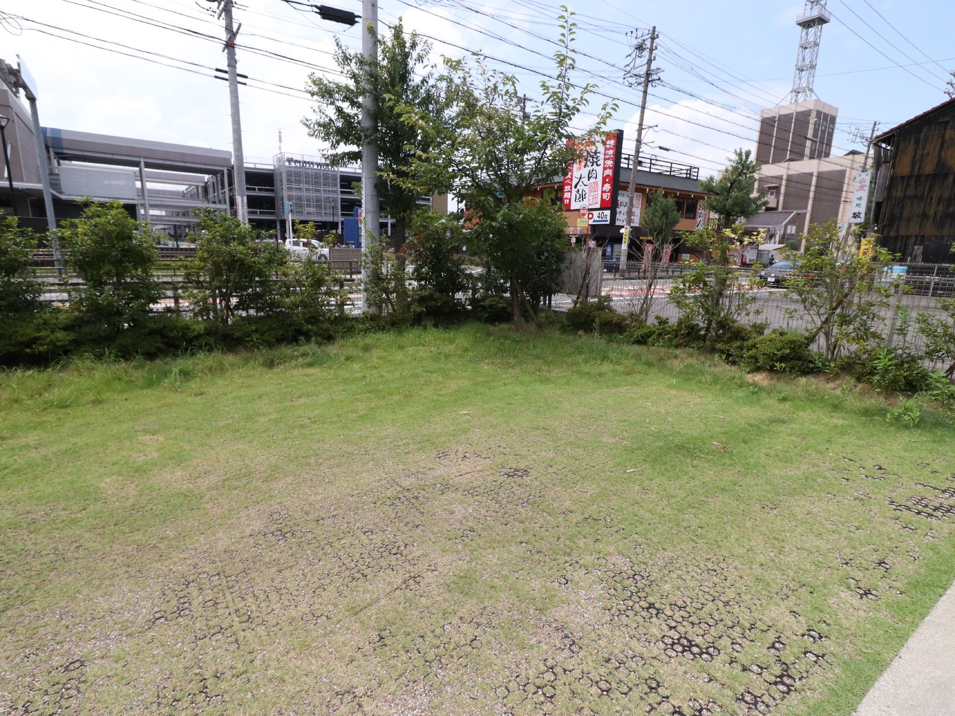 施設周辺の風景が見える。フェンスで囲まれた施設の敷地内は、一部低木と芝生が敷いてある。道路の向こうに車が停まっている。