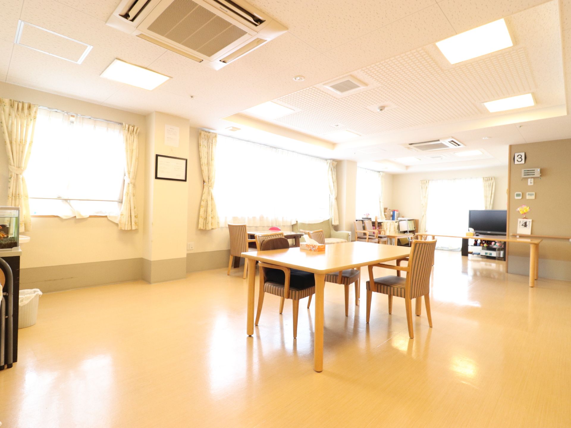 窓にカーテンが取り付けてある部屋で、窓際に一台テレビが置いてある。テーブルには、椅子が四つほど設けてある。