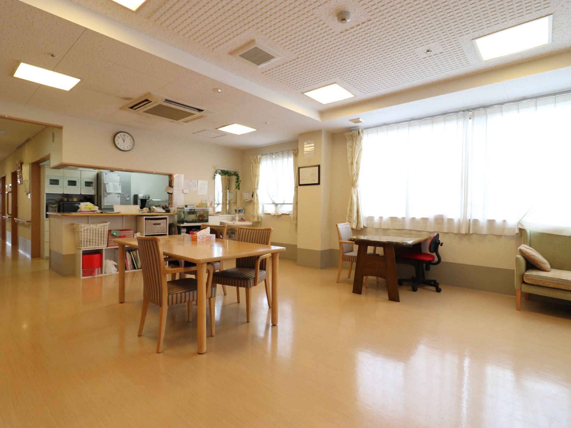 キッチンが見える食堂である。壁掛け時計がある。カウンターの下に収納棚が在り、天板にバスケットが乗っている。