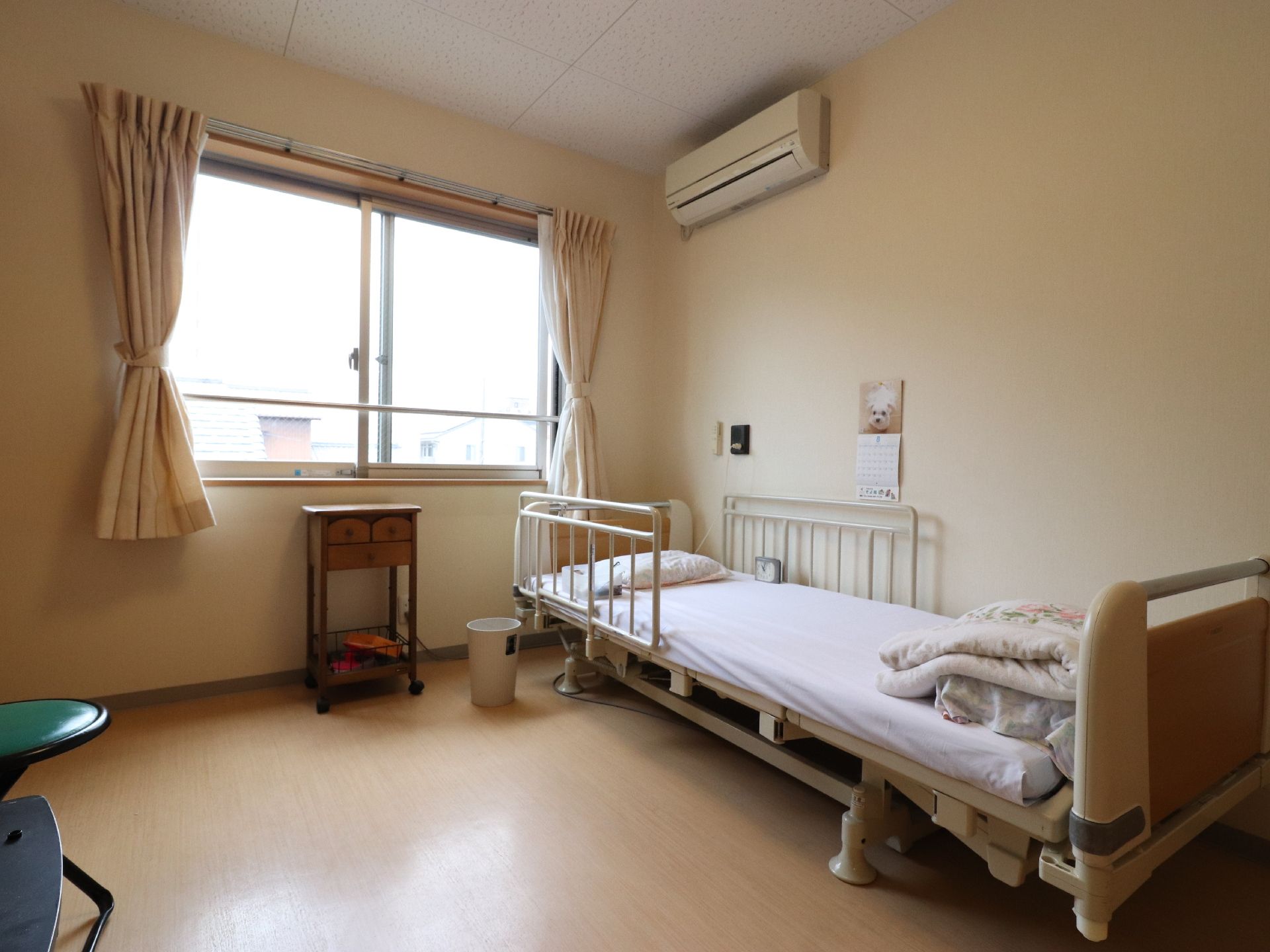 腰高窓のある部屋でカーテンが取り付けてある。布団などの落下防止用の柵が取り付けてある電動ベッドの横の壁にカレンダーが貼ってある。