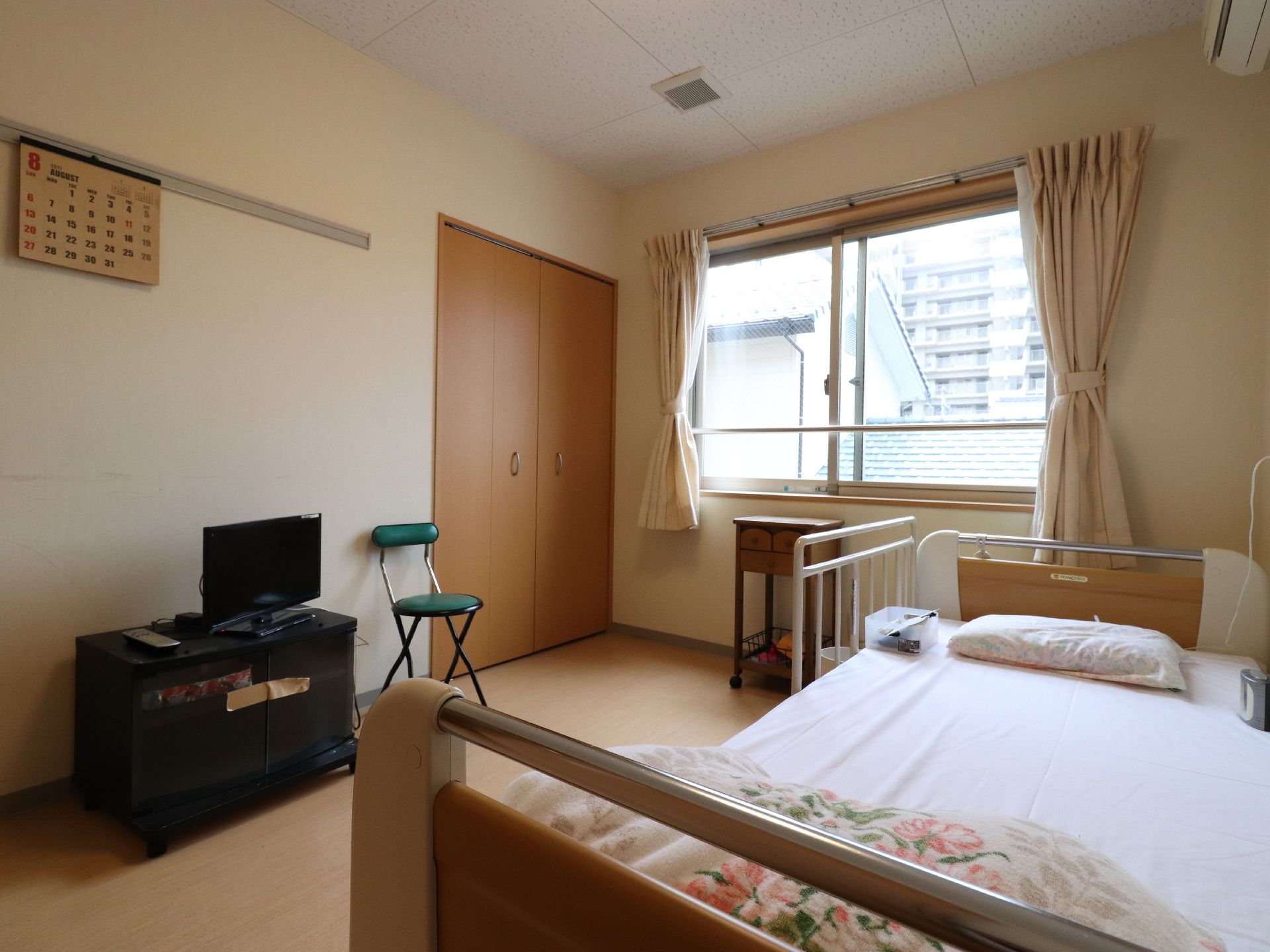 白い壁とフローリングの床の部屋で、壁には月暦が貼ってある。テレビがあり鑑賞できる。座部が緑色の椅子がある。