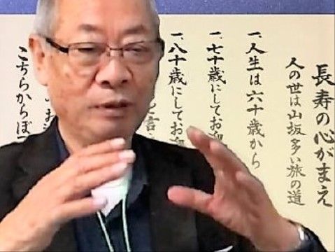 長寿の心がまえと書かれた格言の紙の前で話をする男性