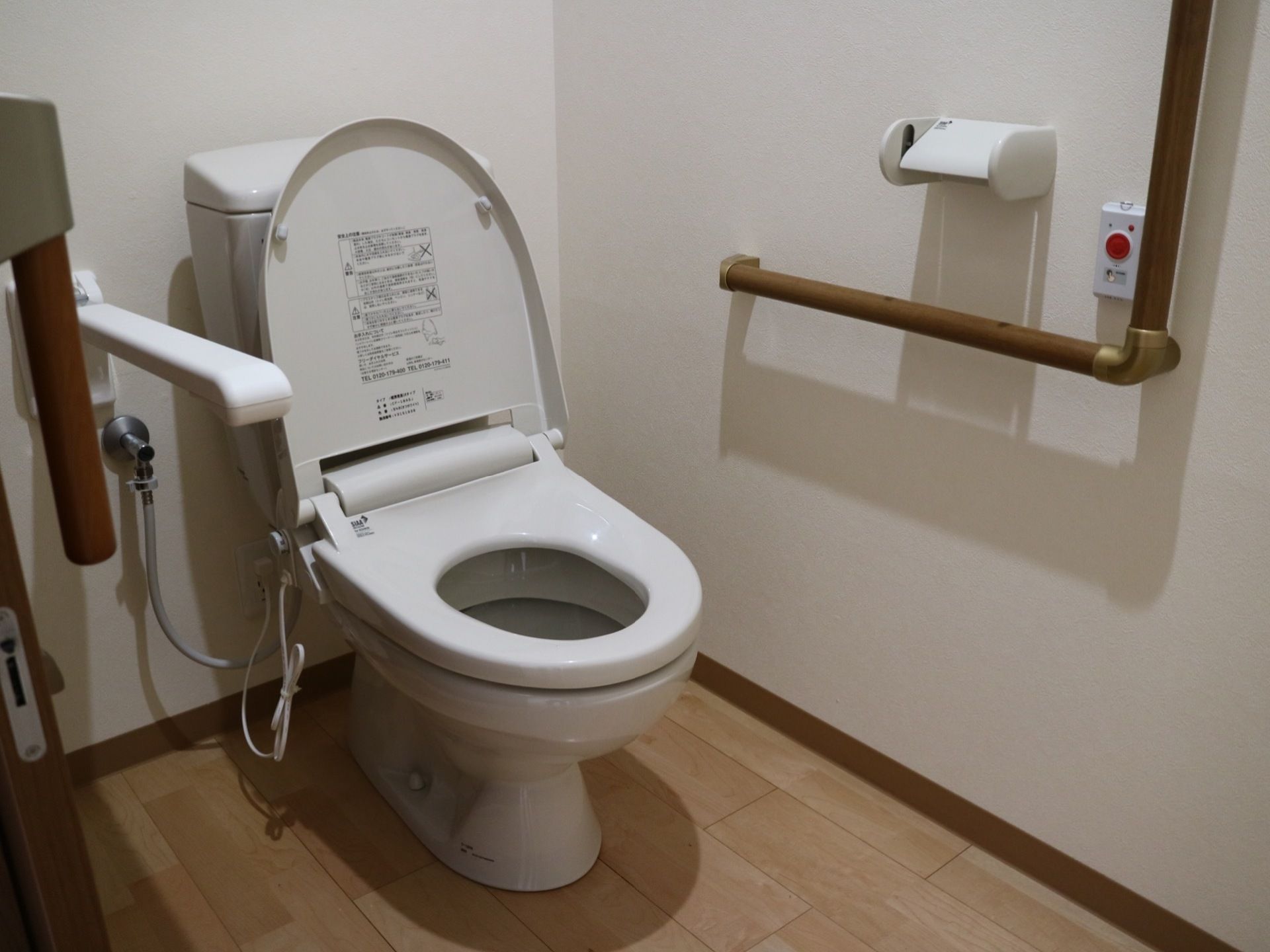 木目調の床で清潔感のあるトイレは、便座の脇に手すりが備わっている。壁に緊急呼び出しボタンが設置されている。