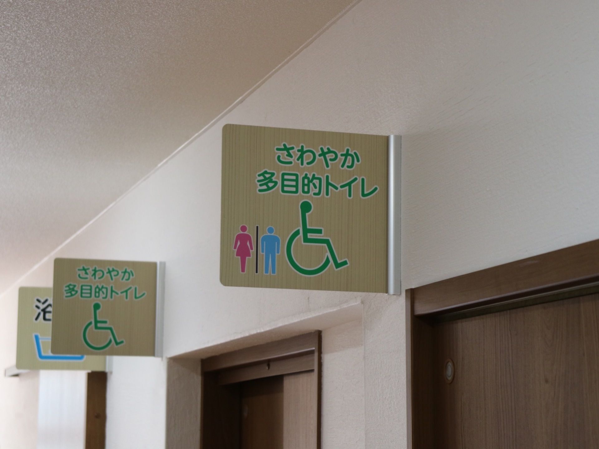 多目的トイレを表示する木目調の看板が壁に設置されている。マークは分かりやすく色が付けられており、見やすくなっている。