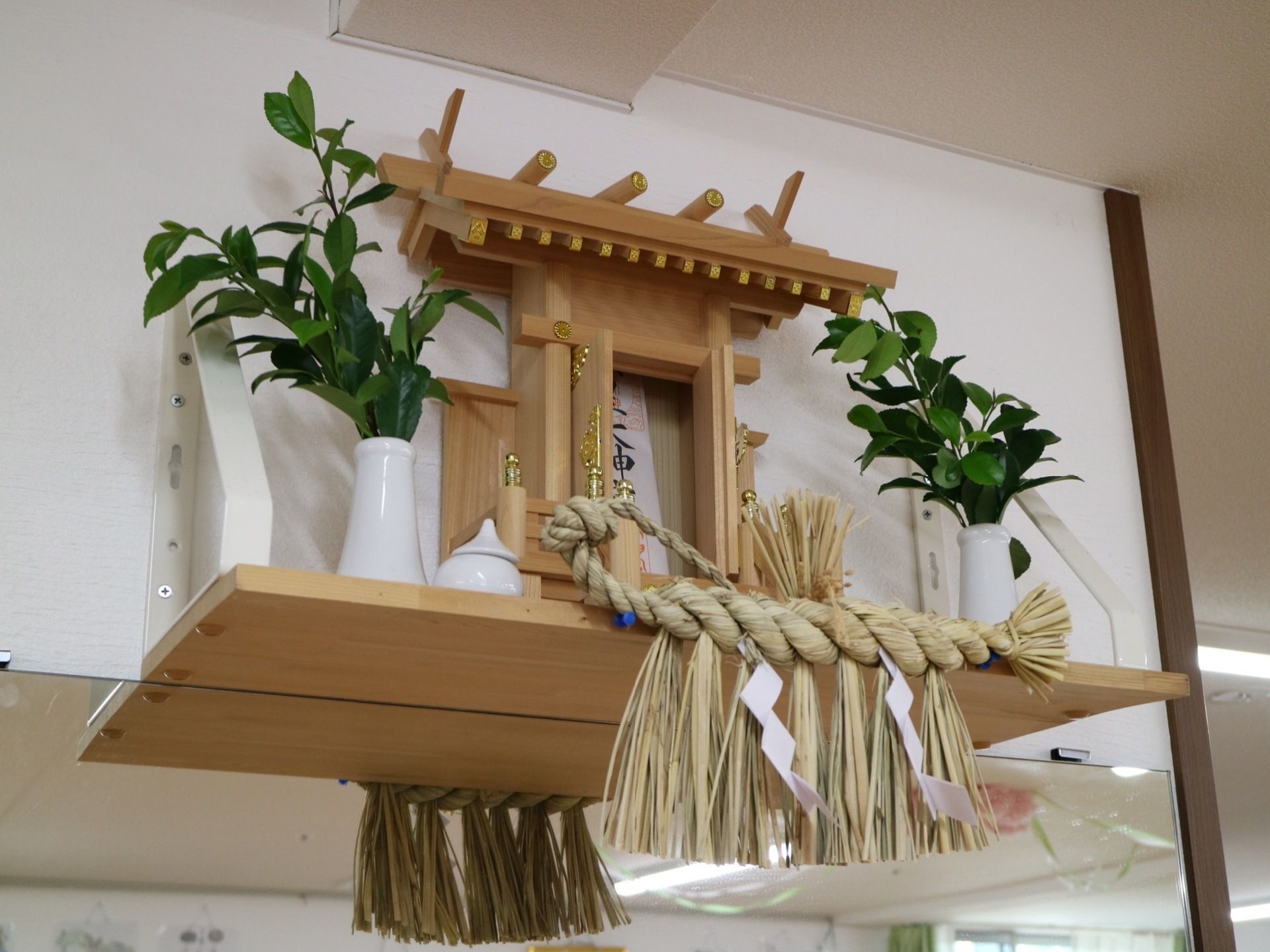 鏡の上の白い壁の部分に、木で作られた神棚が飾られている。神棚には御札とお酒、植物などがお供えされている。