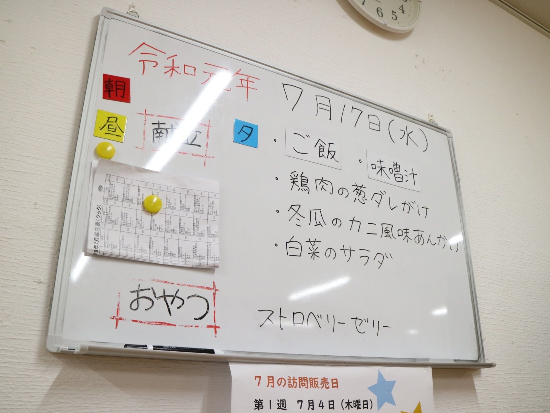 ホワイトボードに一日の献立が昼食、おやつに分けて書かれている。ホワイトボード下には訪問販売のお知らせが掲示されている。