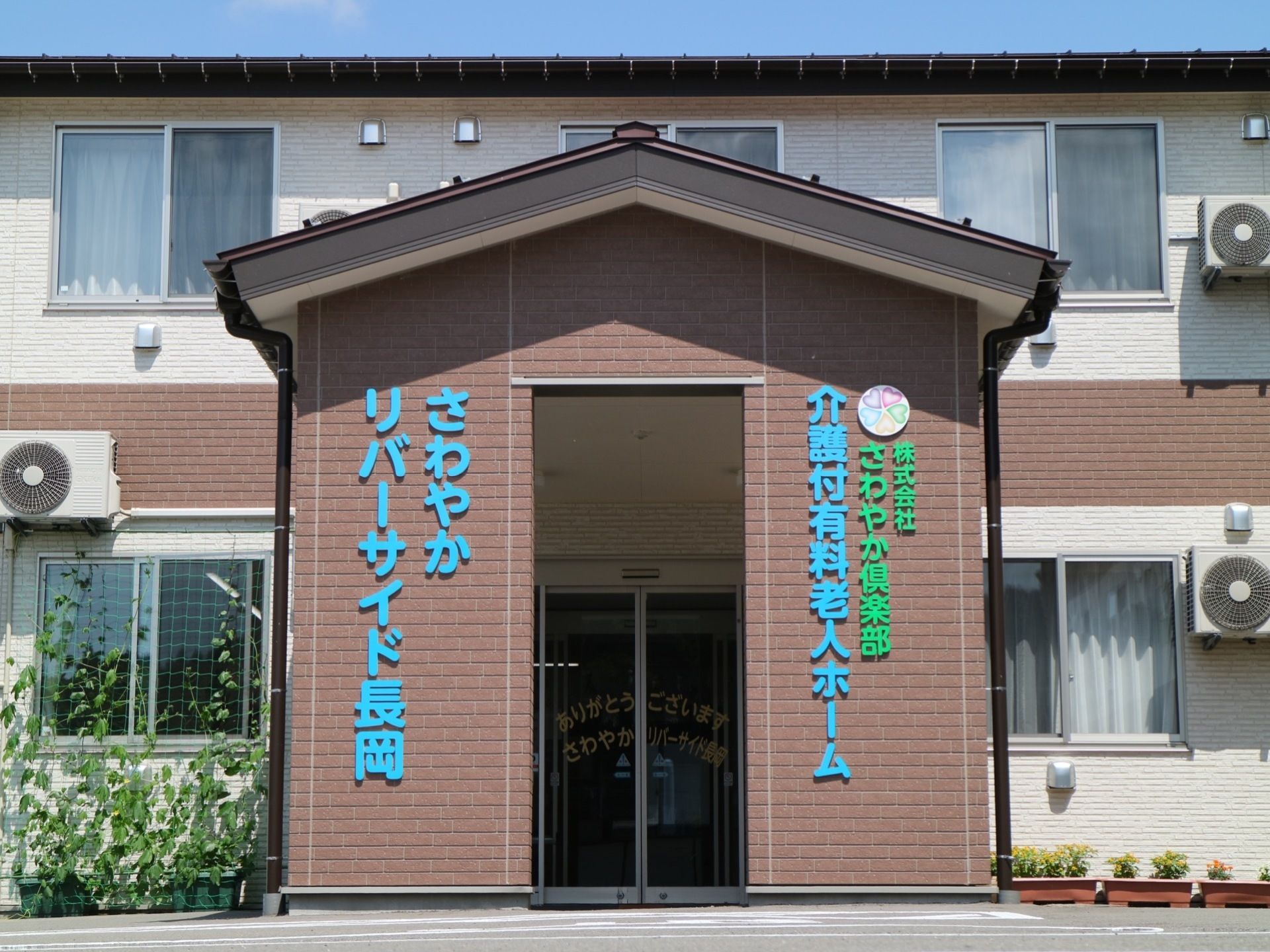 白とブラウンを基調とした3階建ての施設。施設名が水色と緑色の文字で分かりやすく掲示されている。玄関前は屋根が設置されている。