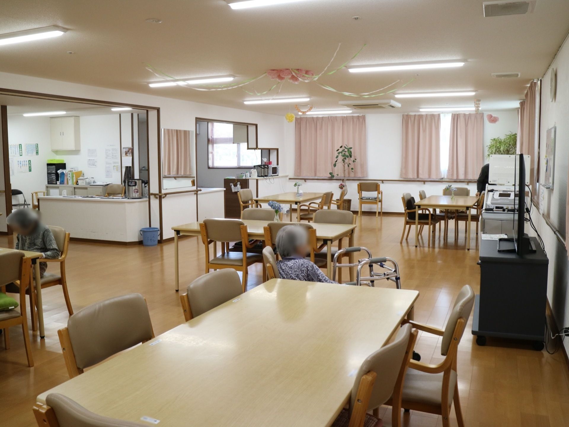 ダイニングテーブルと椅子が置かれた共有スペース。テレビがあり、入居者様がテレビ鑑賞をしている。奥に調理スペースがある。