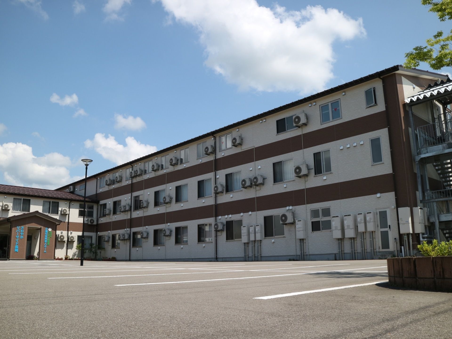 ３階建ての施設に非常用階段が設置されている。広々とした駐車場があり、駐車スペースが白線で区切られている。