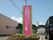 施設横の花壇に看板が建てられている。看板はピンク色で緑色のラインで縁取られており、施設のマークと施設名が大きく描かれている。