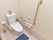 洋式トイレの床には転倒防止用のマットが敷かれている。壁には手すりと呼び出しボタンが設置されている。タンクの上に花が飾られている。