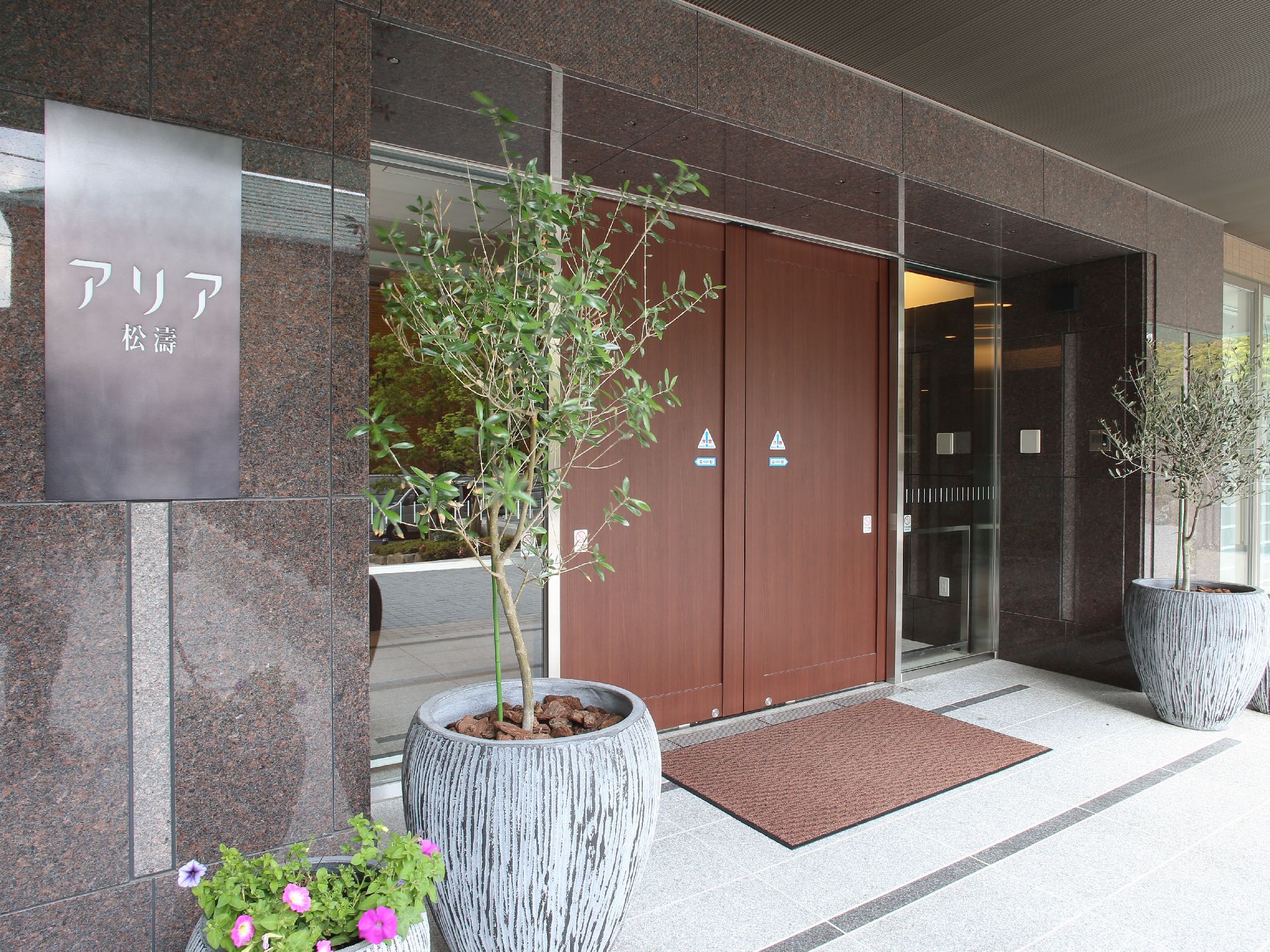 インパクトのある大きな陶器鉢に木が植えられている。ブラウンの自動ドアが設置されており、出入り口には玄関マットが敷かれている。
