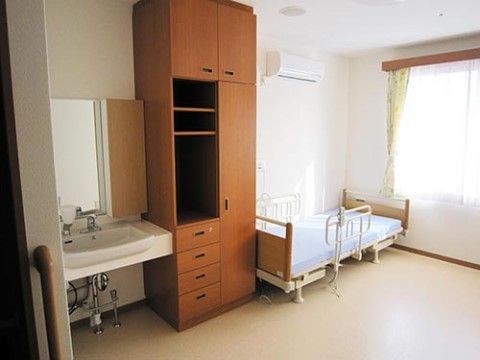 介護用ベッド、チェスト、洗面台、エアコンが設置された広々とした居室である。窓からは日が差し込み、明るい雰囲気である。