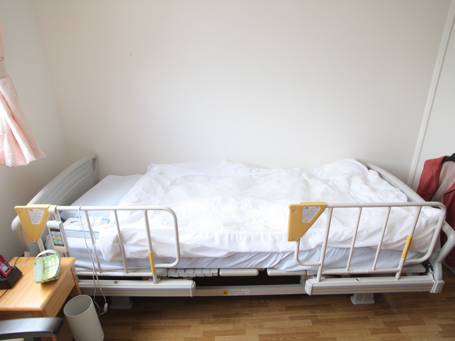 フローリングの居室に介護ベッドが置かれている。白い壁で明るい空間になっており、窓がある場所にはピンクのカーテンが付いている。