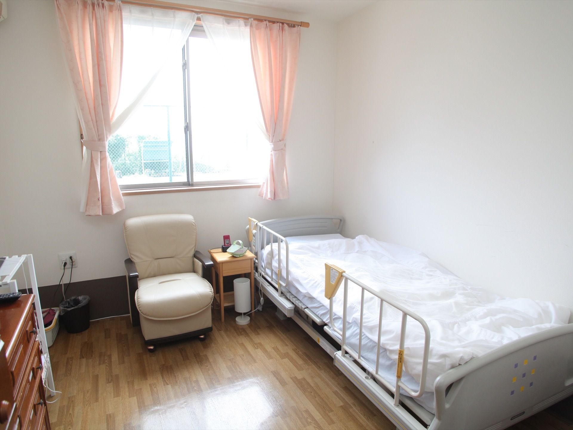 窓から光が入る、バリアフリーの明るい居室である。ベッドやサイドテーブル、一人掛けのソファなどが置かれている。