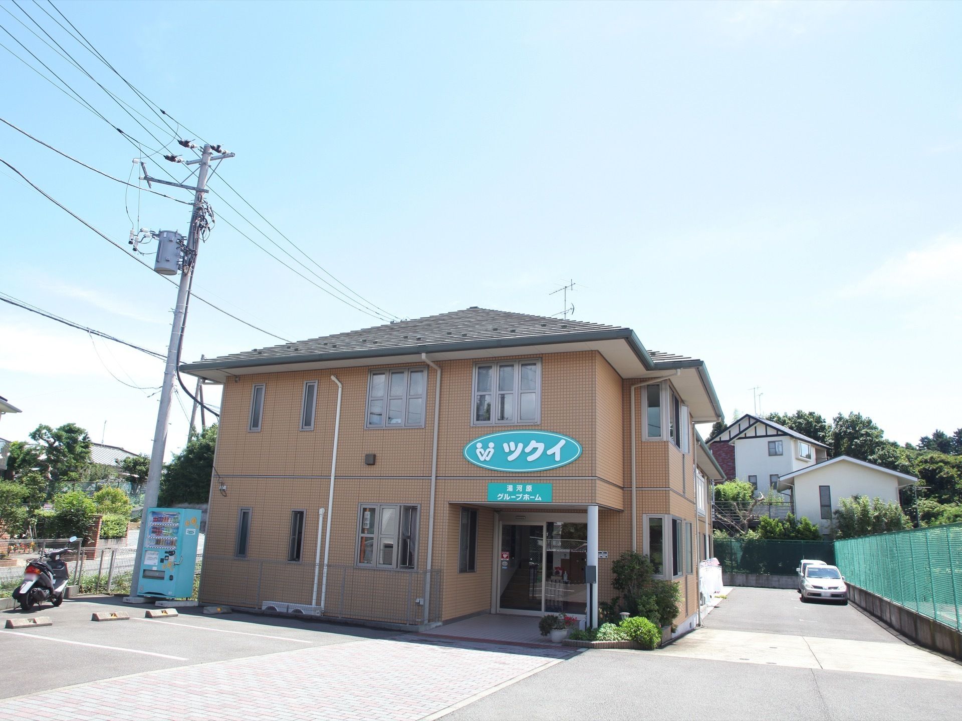 玄関の側には植物が植えられている。建物の横には自動販売機が設置されていて、駐車場にはバイクが停められている。