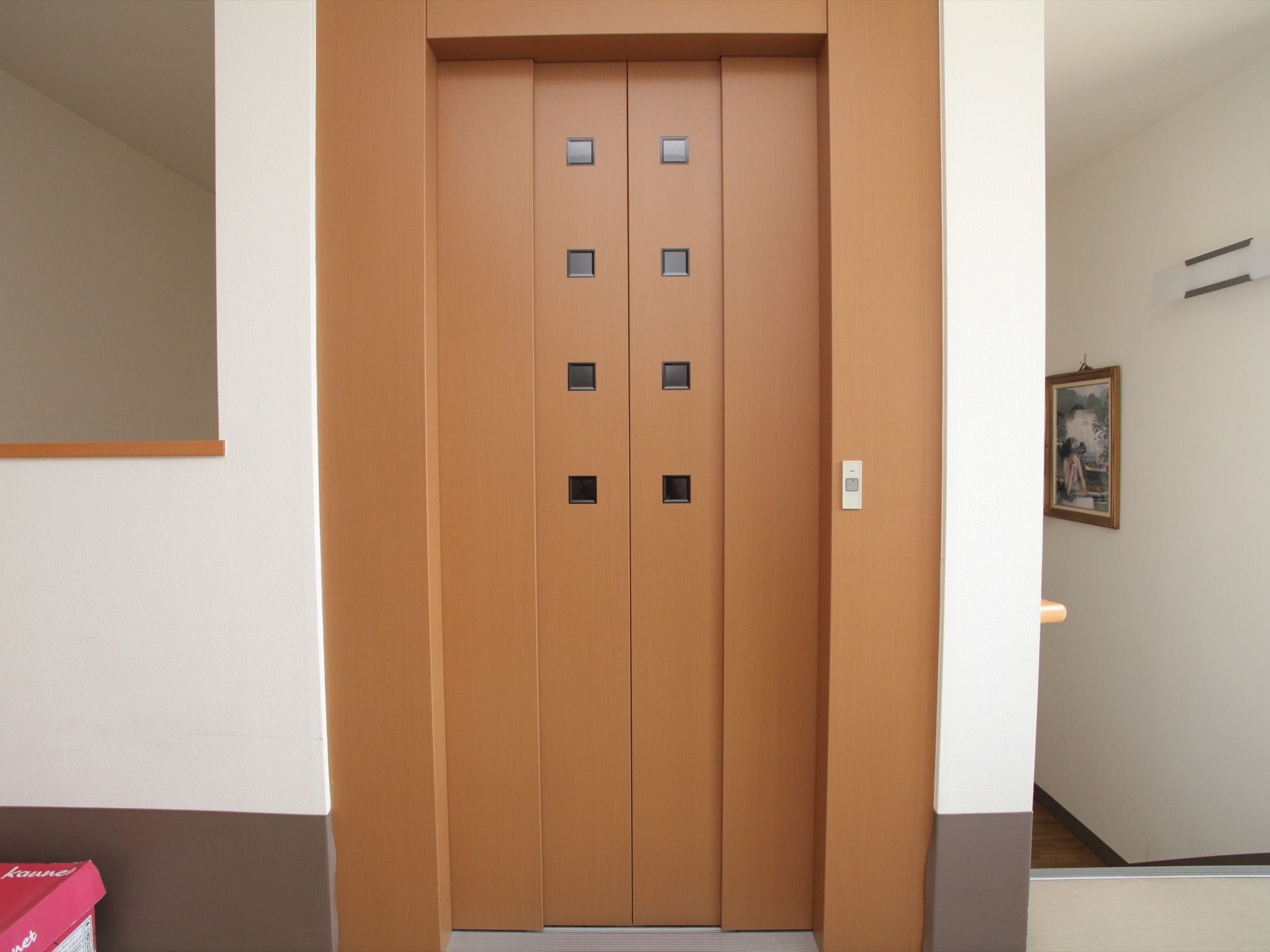 エレベーターのドアは茶色で、四角い小さなガラスがはめられている。横の壁には小さなボタンが付いている。