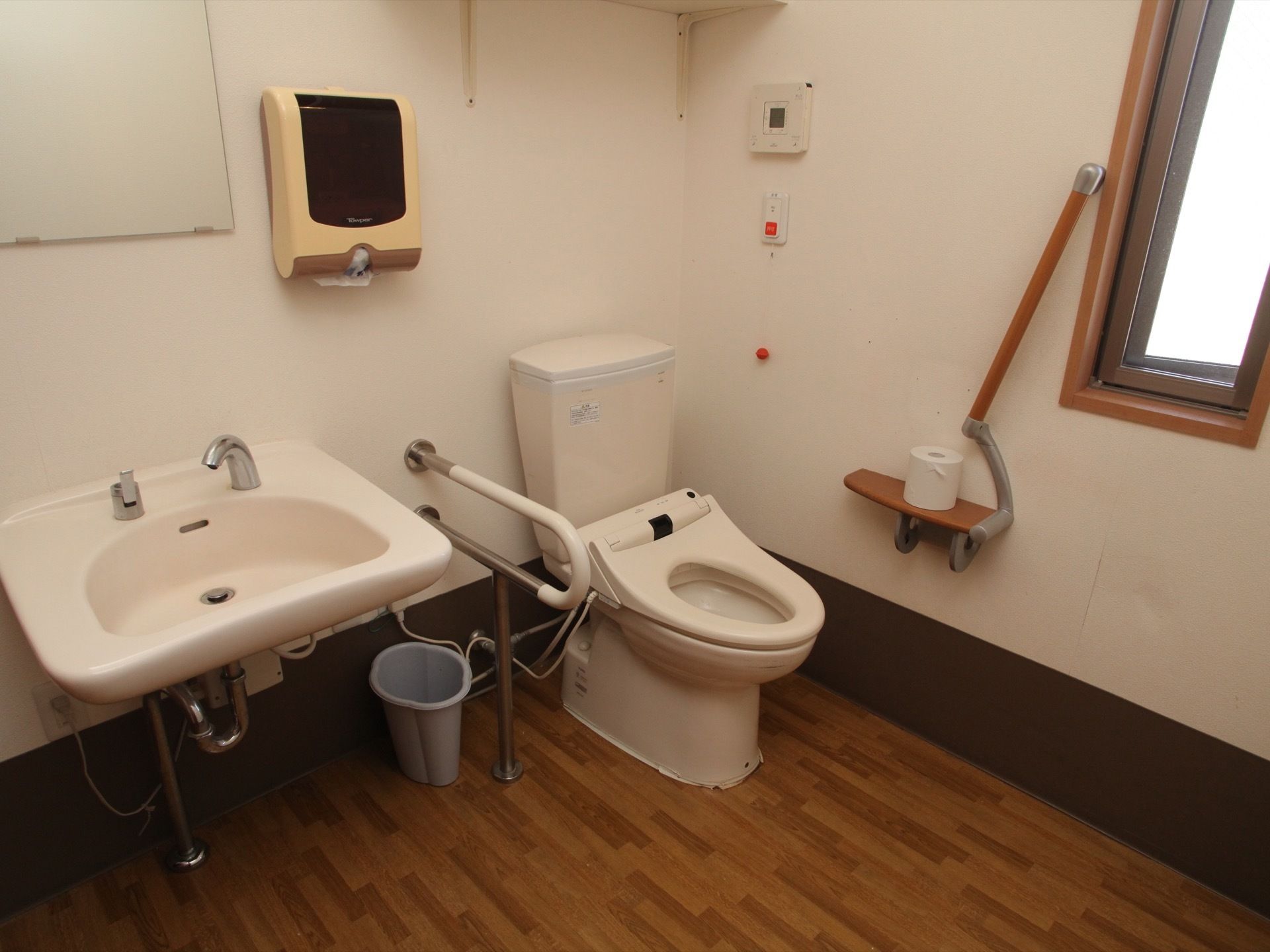 床がフローリングになった広いトイレである。便座の横の壁には手すりが斜めに取り付けられており、その下の台にはトイレットペーパーが置かれている。