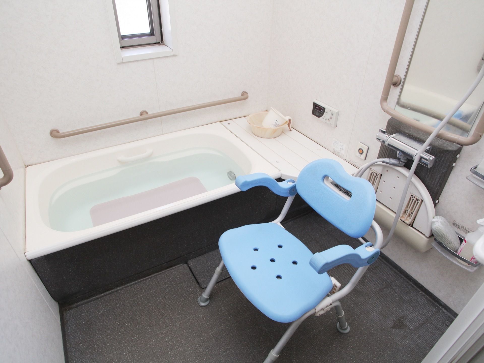 床がグレーで壁が白い、清潔感のあるユニットバスである。ブルーのシャワーチェアが置かれ、壁には手すりや呼び出しボタンなどが設置されている。