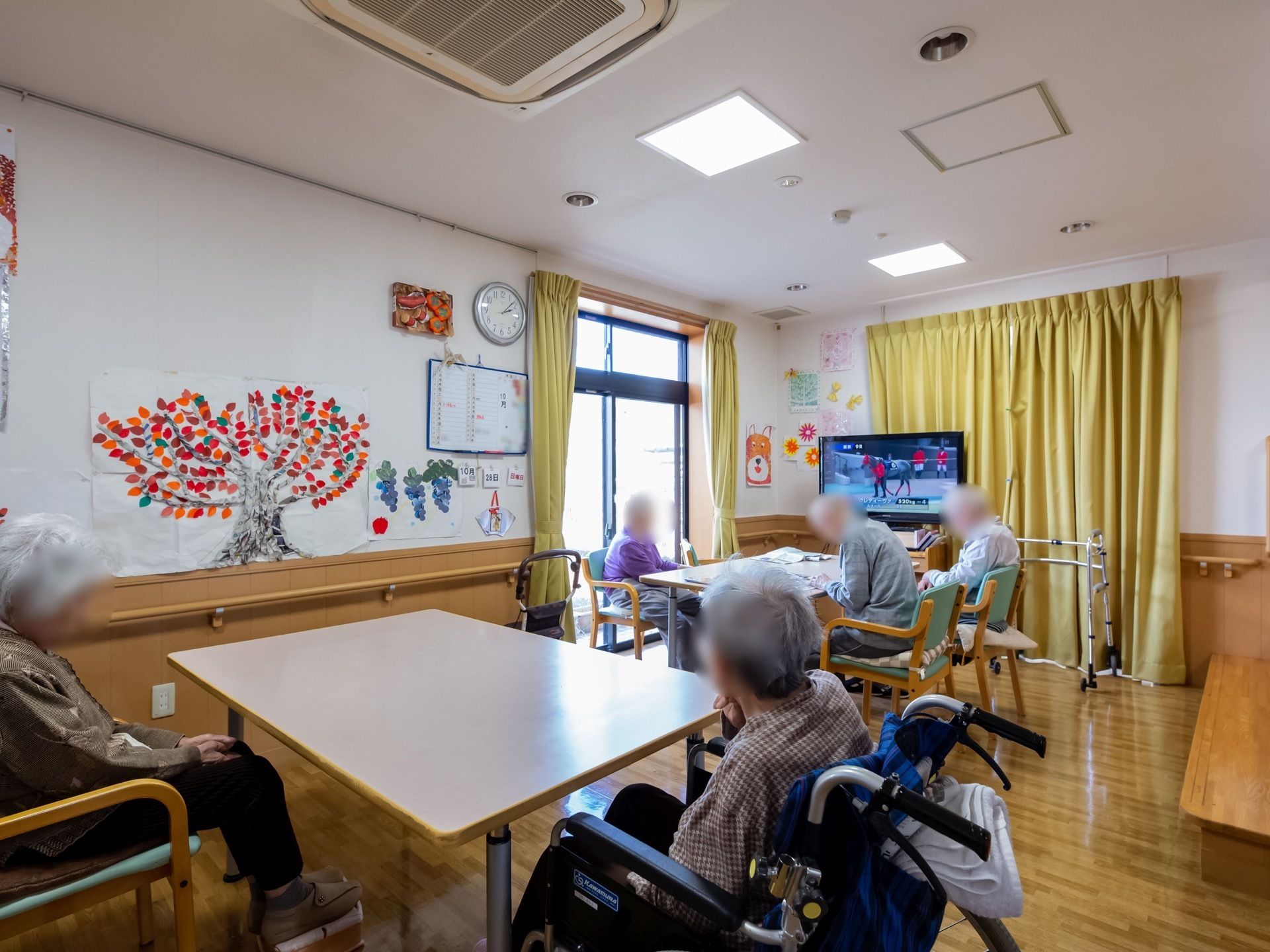 空調のついた広い空間に、テーブルが並んでいる。入居者様が5人ほど腰を掛けており、テレビを鑑賞している。