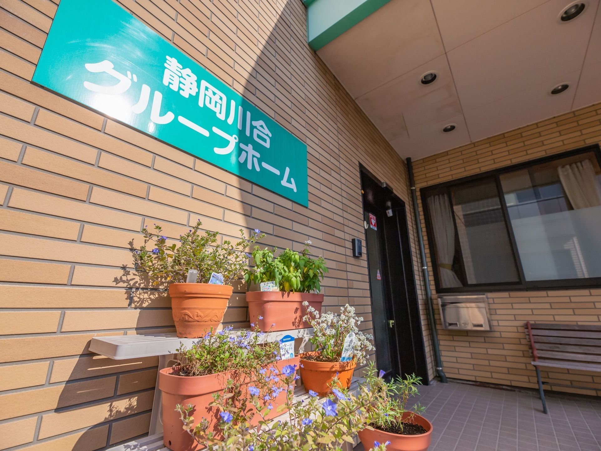 茶色いタイルの壁の前にひな壇があり、植木鉢の花が並べられている。その上にはホームの看板が掛けられている。