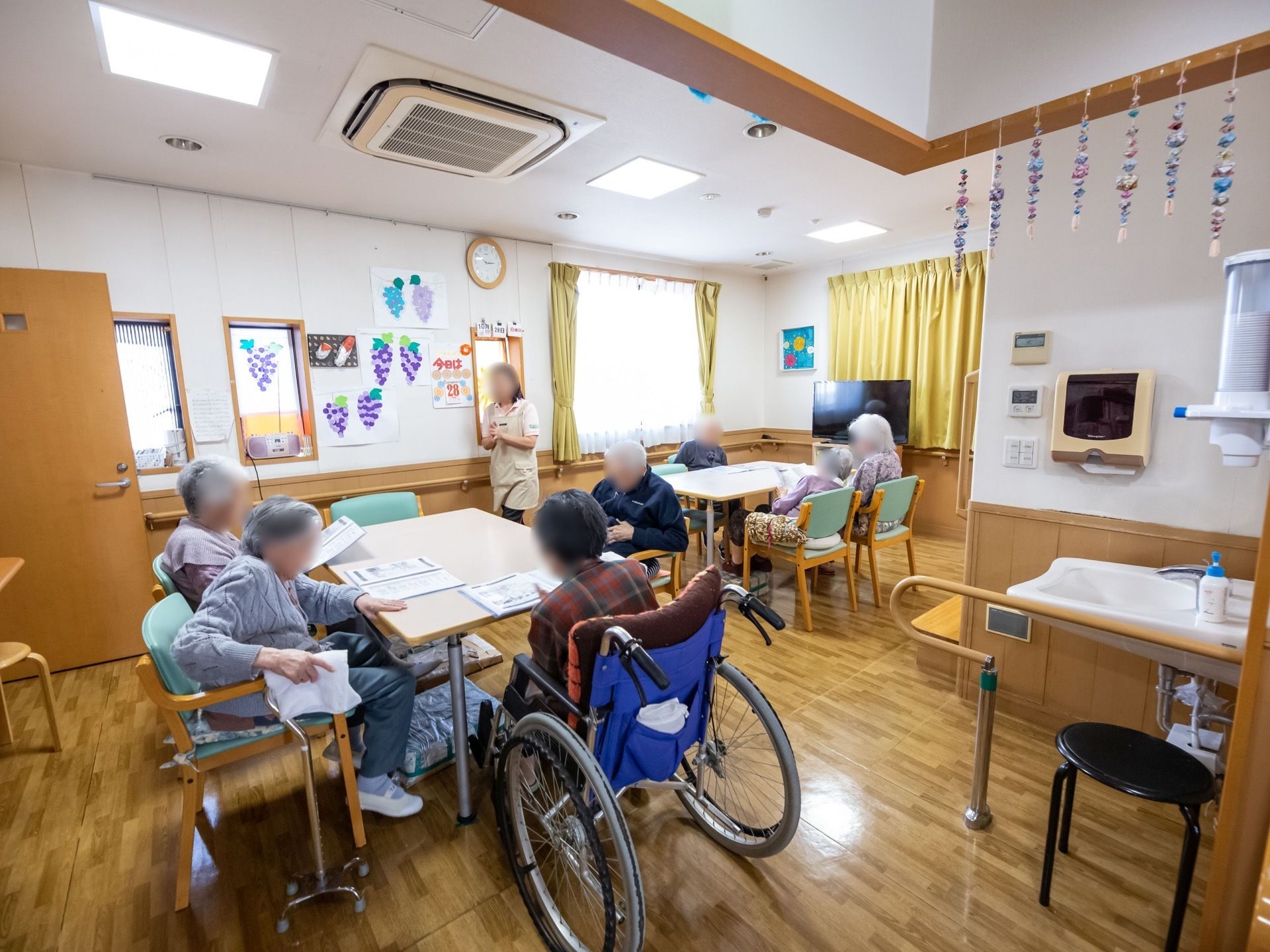 入居者様が集まってテーブルについている。女性のスタッフが何かを説明し、入居者様は手元の紙を見ている。